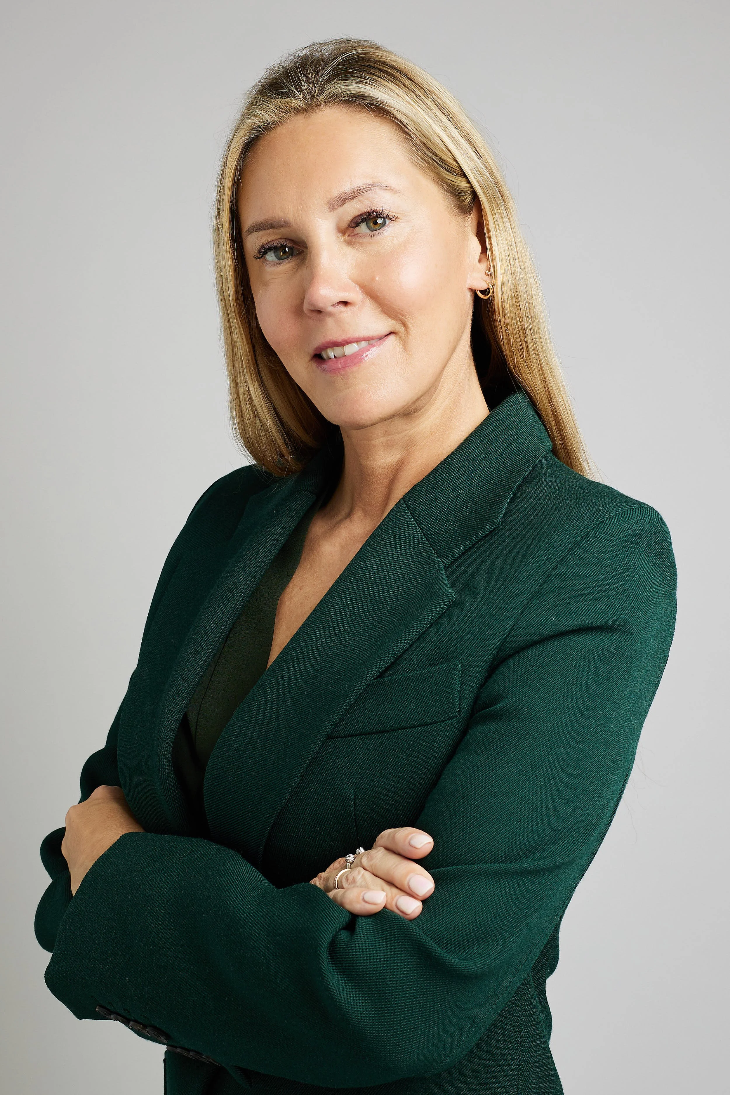 A woman with blonde hair, wearing a dark green blazer, standing with her arms crossed and smiling at the camera against a plain light gray background.