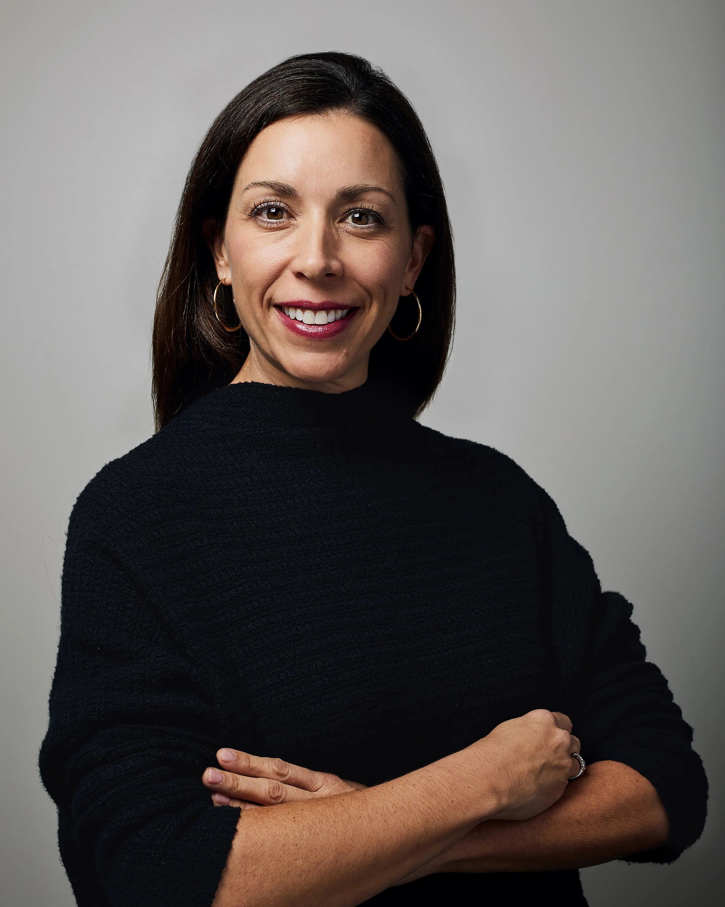 A woman with shoulder-length dark hair smiling, wearing a black sweater and gold hoop earrings, standing with arms crossed against a gray background.