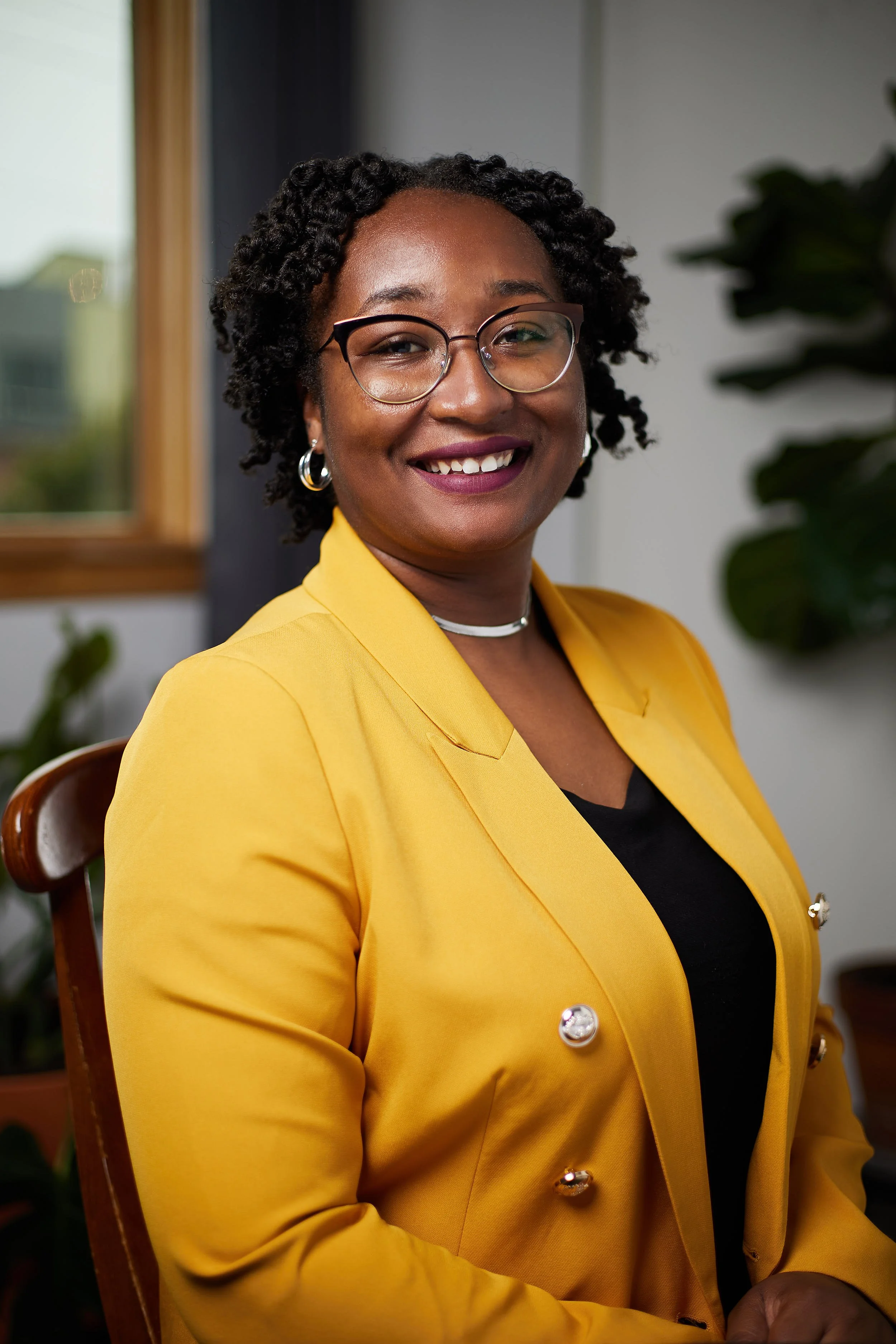 A woman with curly black hair and glasses, smiling, dressed in a yellow blazer over a black top, sitting in a room with plants and a window in the background.