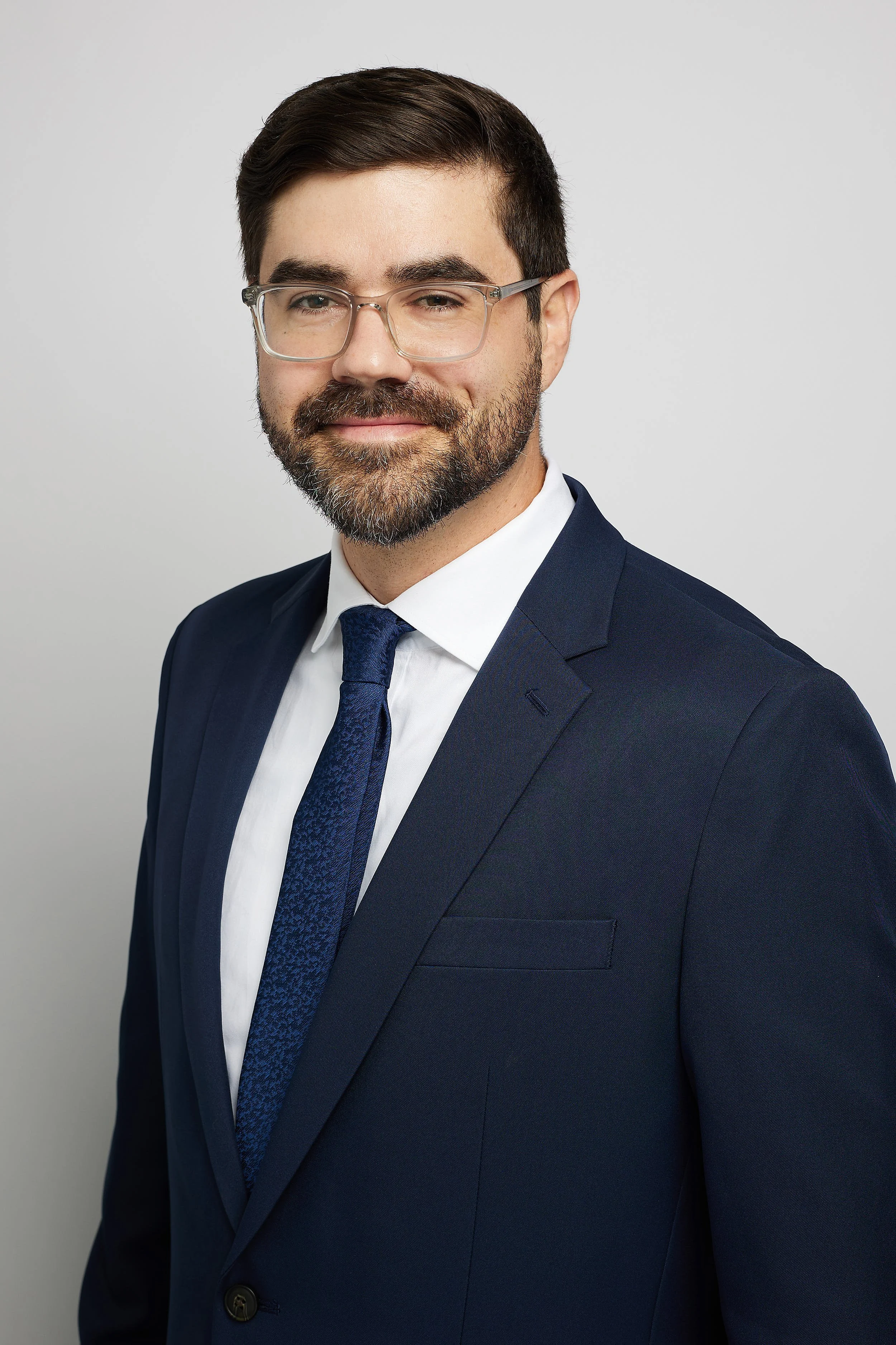 A professional man with glasses, dark hair, and a beard, wearing a navy suit, white shirt, and navy tie, standing against a plain light gray background.