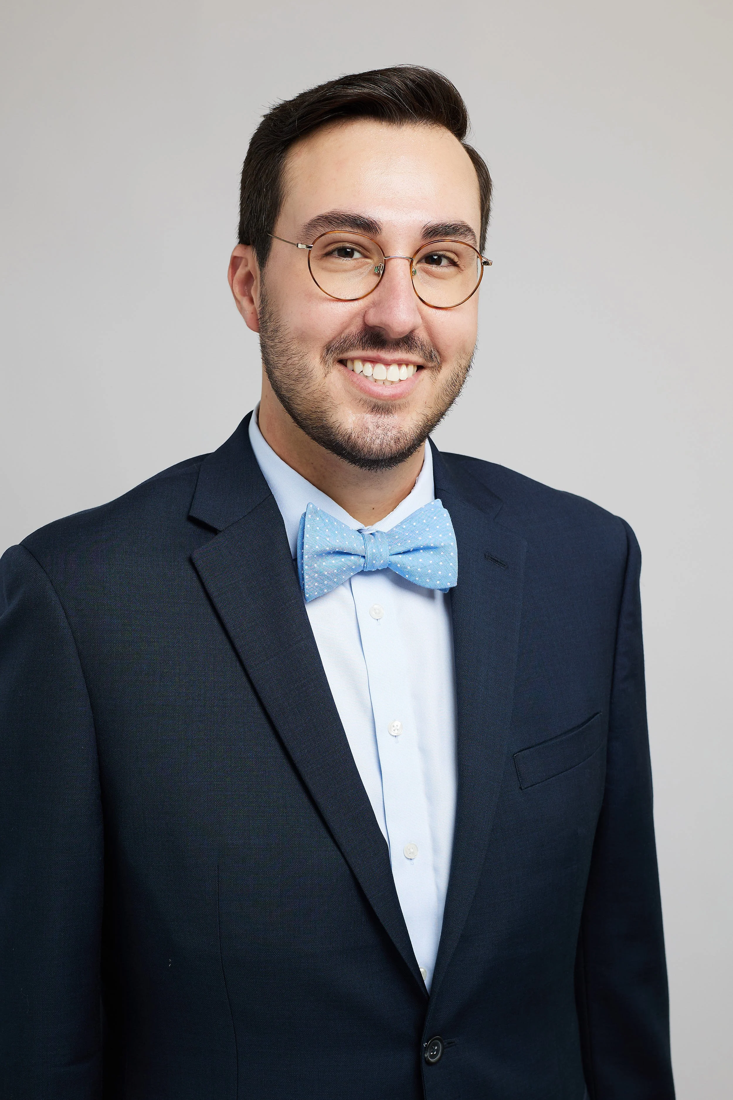 A man in a formal dark suit, light blue dress shirt, and light blue bow tie, smiling and wearing glasses, posing against a plain light background.