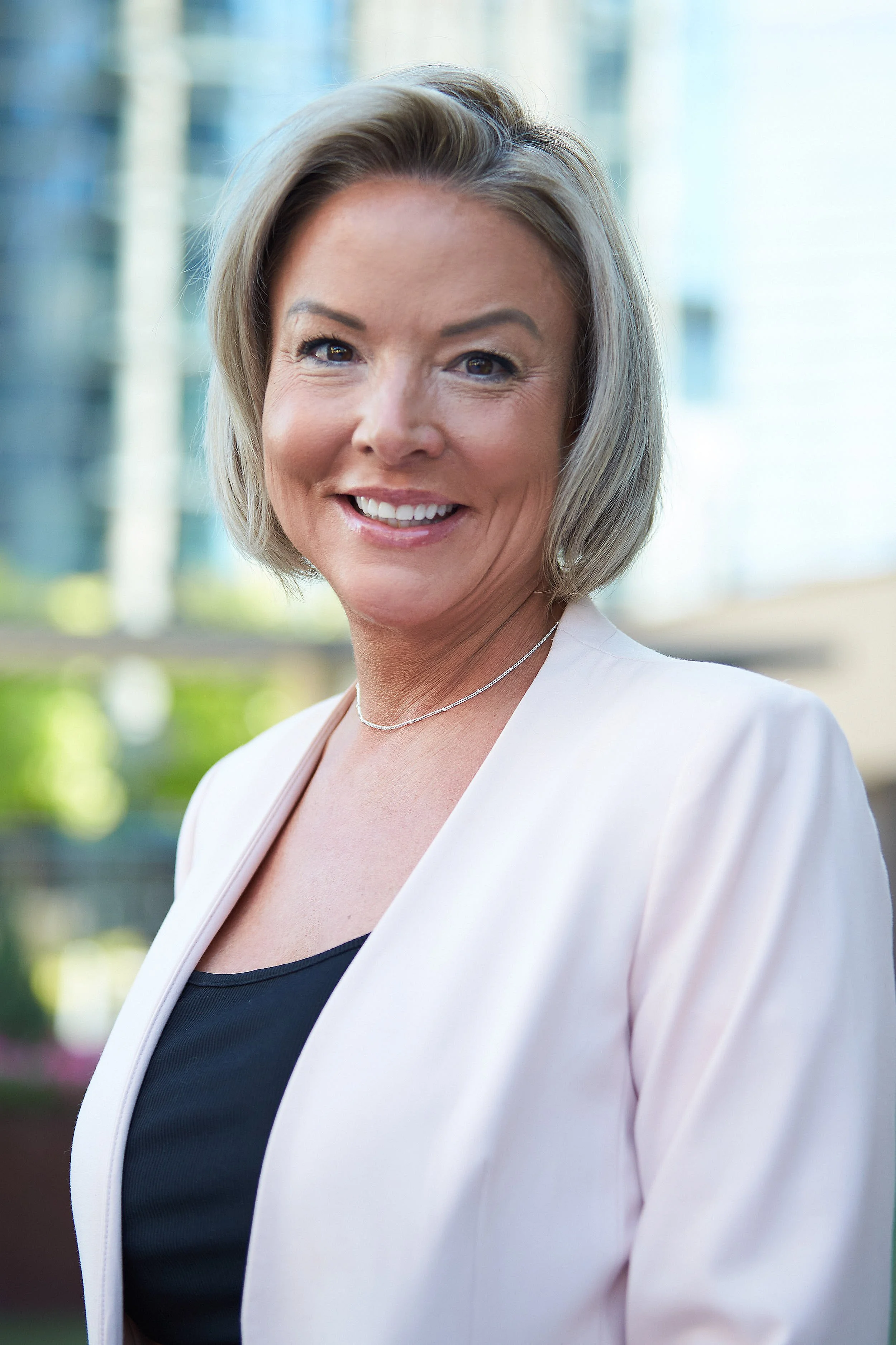 A woman with short blonde hair wearing a white blazer and black top standing outdoors with blurred city buildings in the background, smiling at the camera.