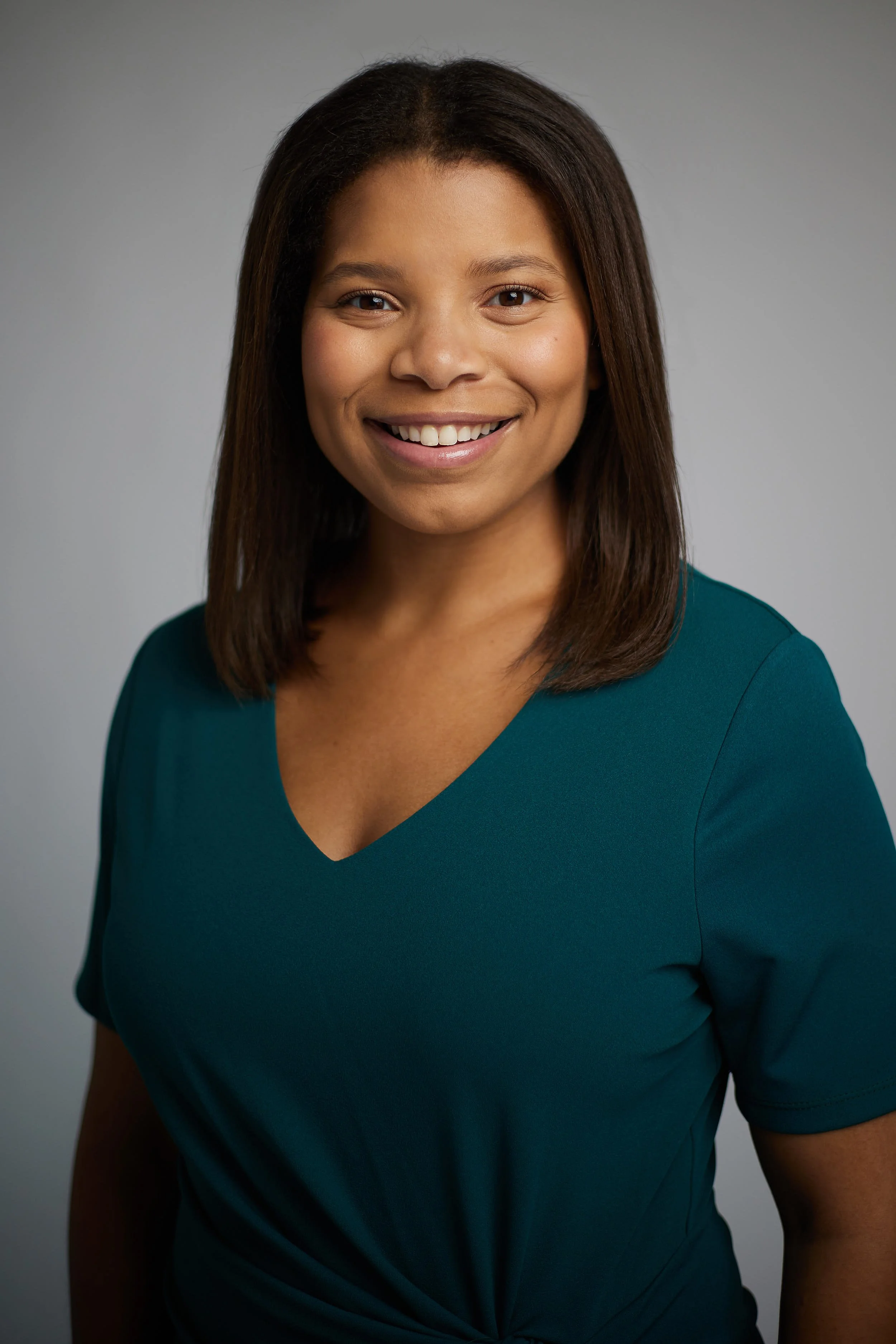 A woman with medium-length brown hair smiling, wearing a teal V-neck shirt, standing against a plain gray background.