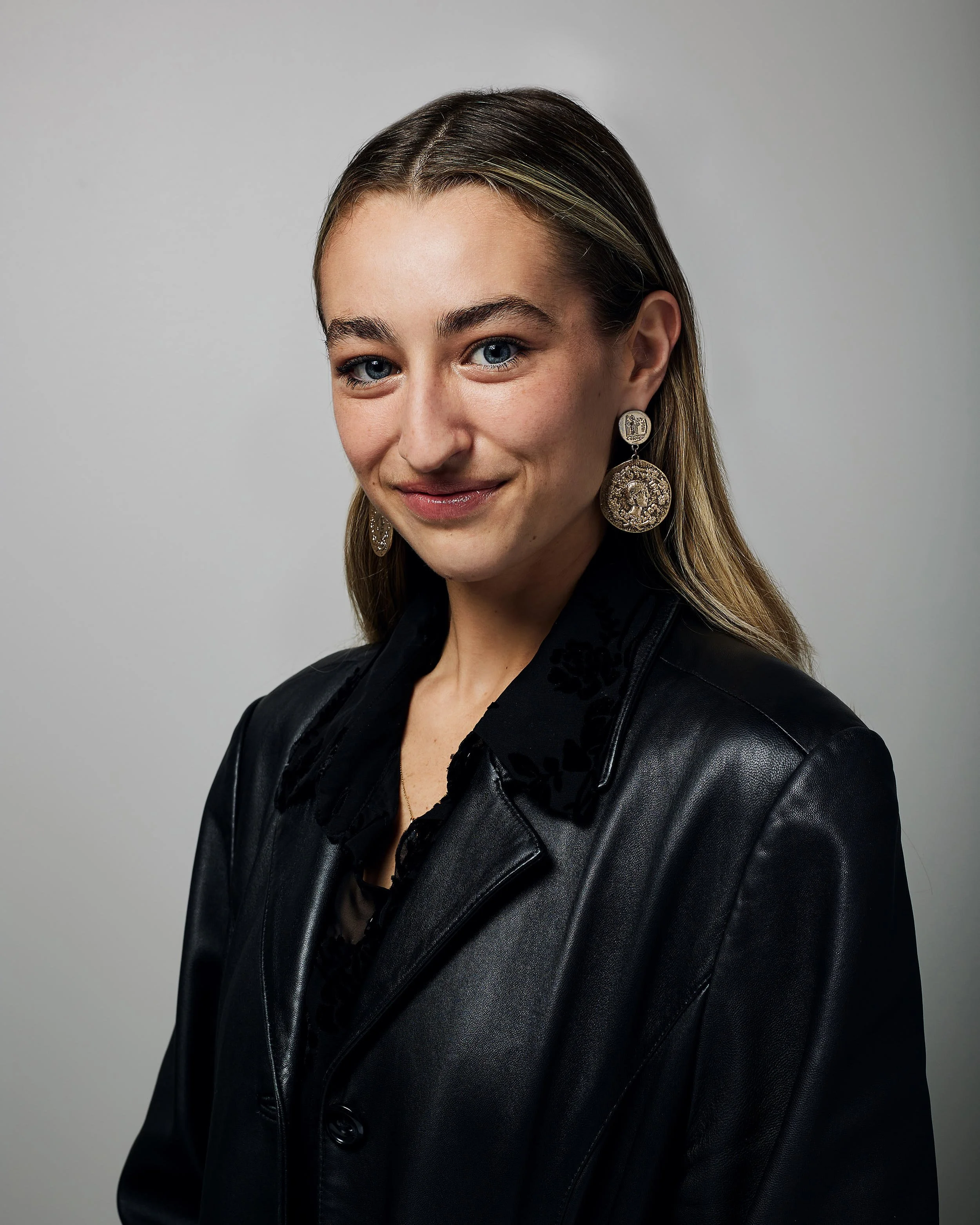 A woman with long hair wearing a black leather jacket and large earrings, smiling against a plain gray background.