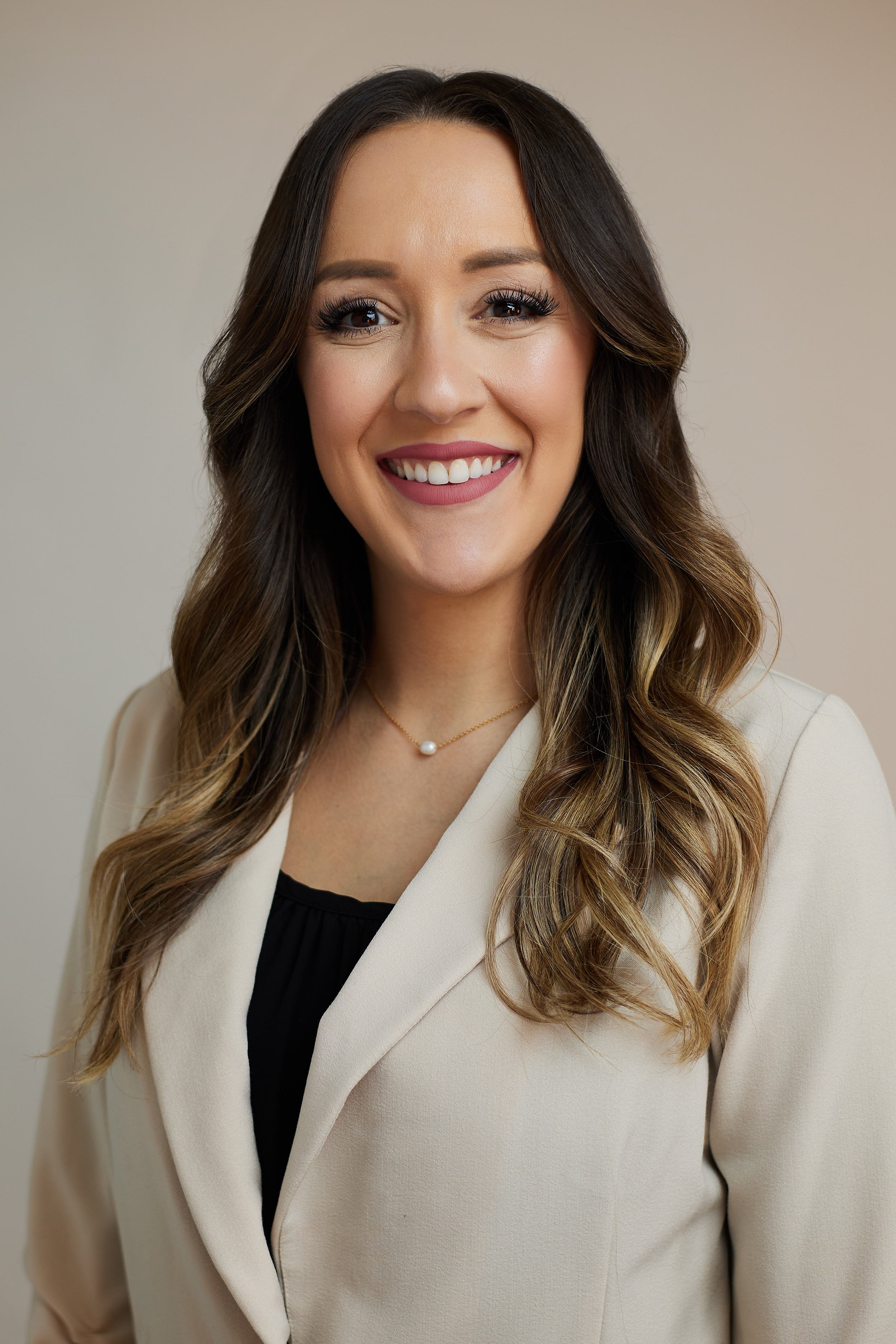 Professional woman with long wavy brown hair, wearing a beige blazer, black top, and pearl necklace, smiling against a neutral background.