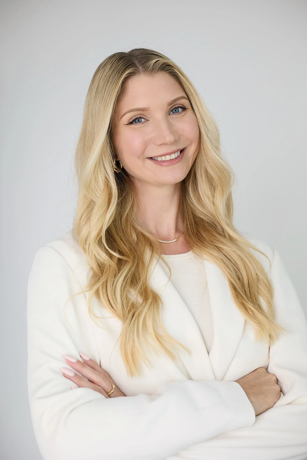 A woman with long blonde hair smiling, wearing a white blazer and jewelry, posing against a light gray background.