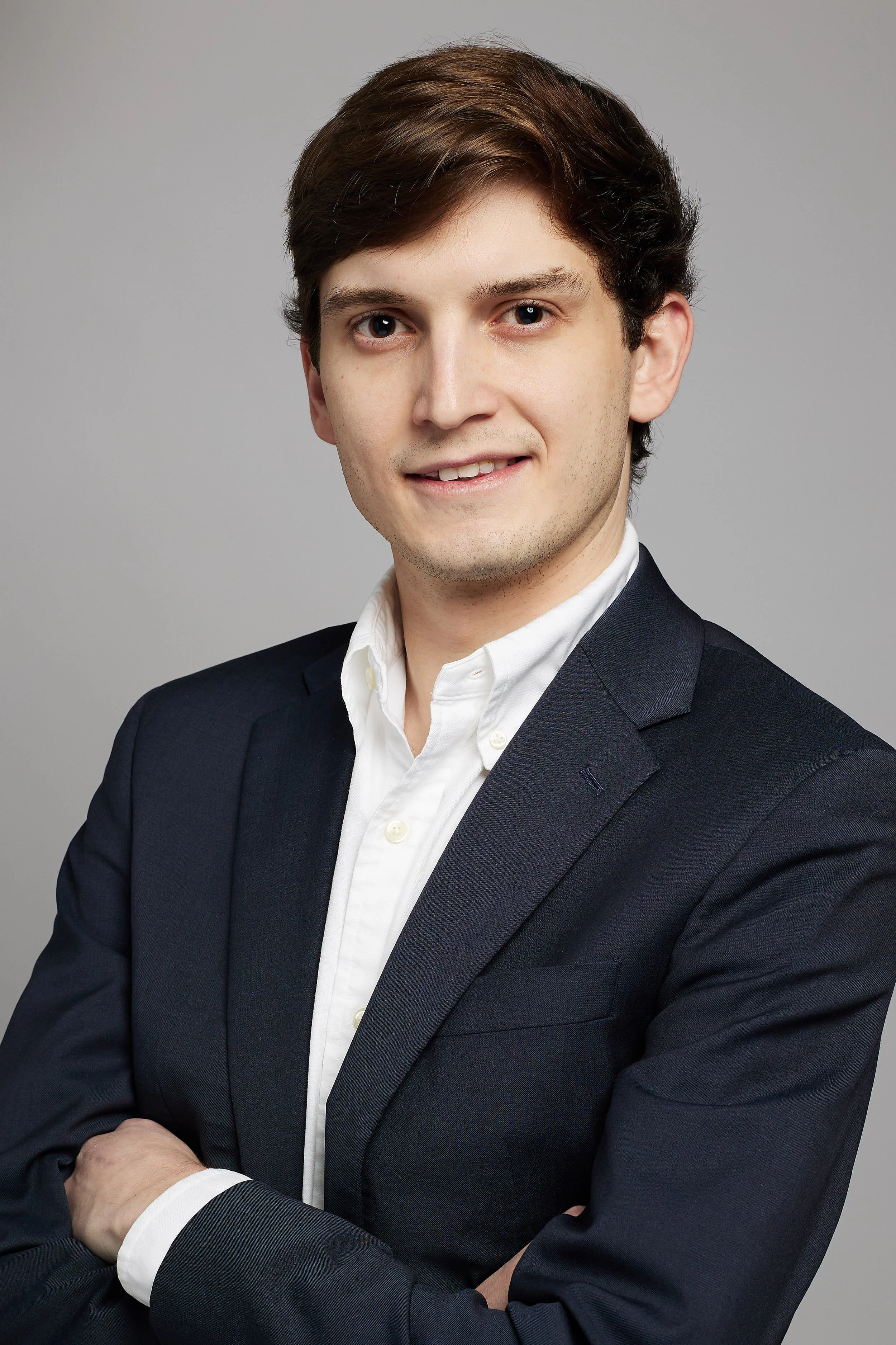 A young man with brown hair wearing a dark suit and white shirt, standing with arms crossed against a plain gray background.