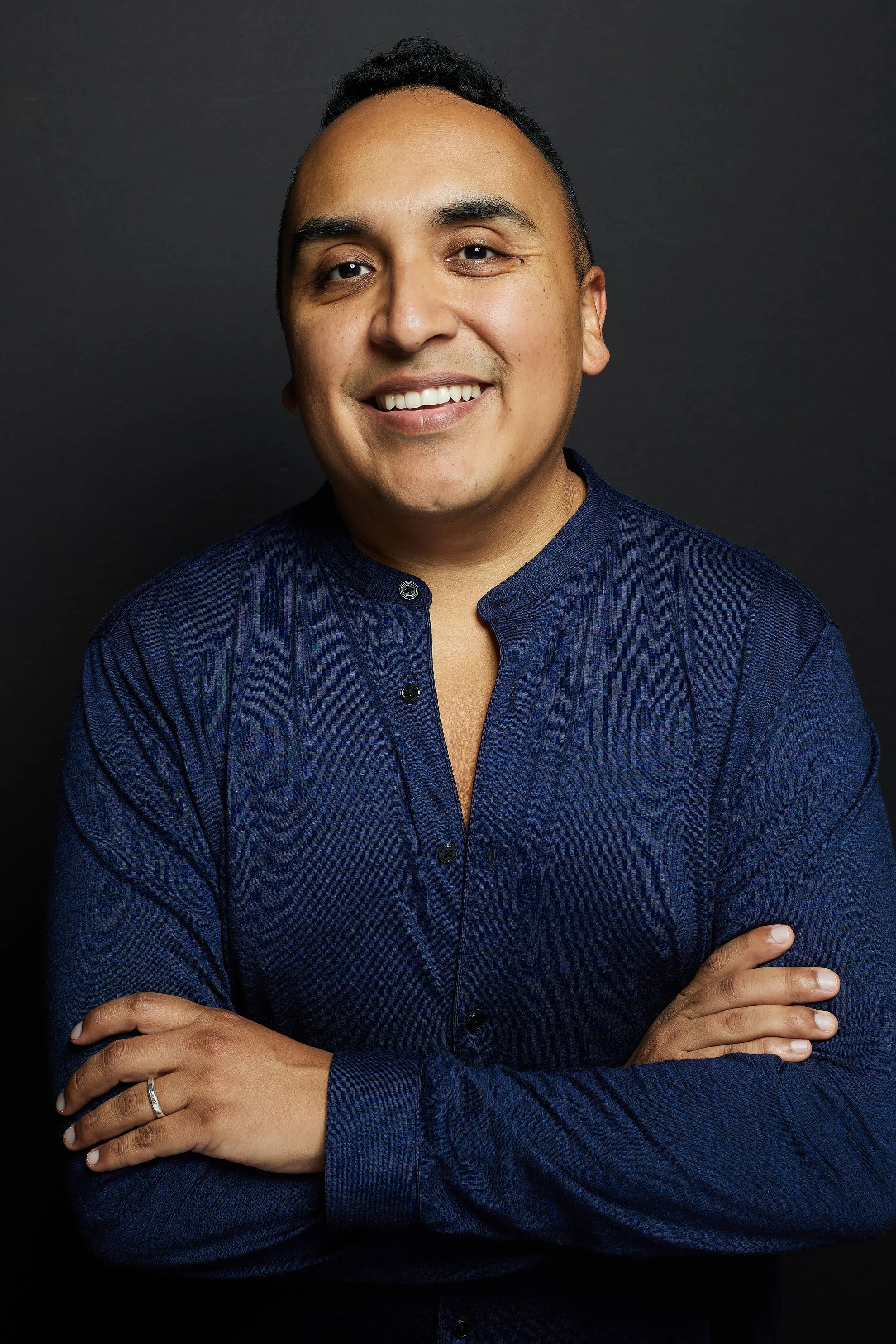 A smiling man with crossed arms, wearing a dark blue shirt, standing against a black background.