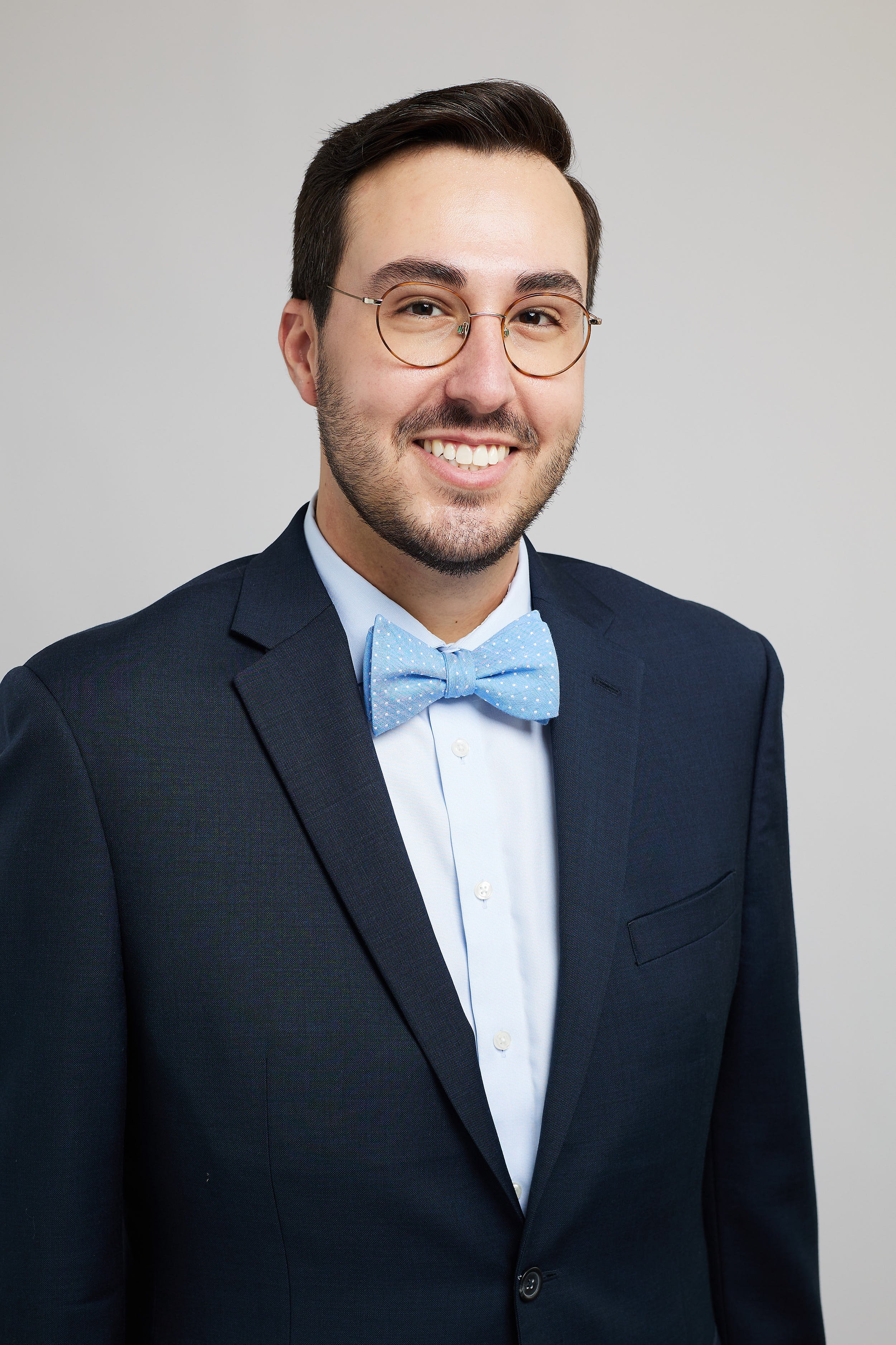 A man in a dark suit with a light blue shirt and matching bow tie, wearing round glasses and smiling against a plain light background.