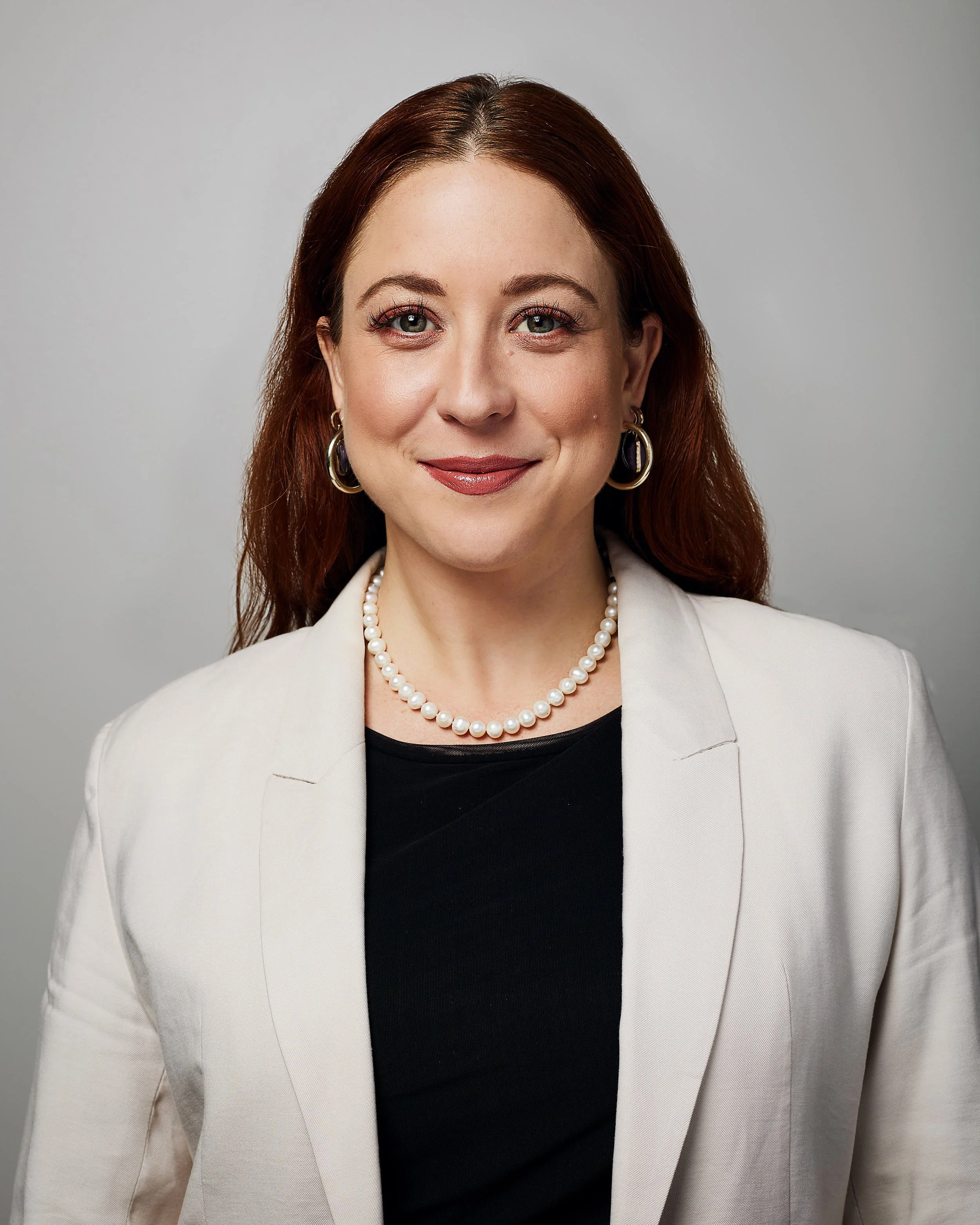 Headshot of young business woman with red hair and pearl necklace.