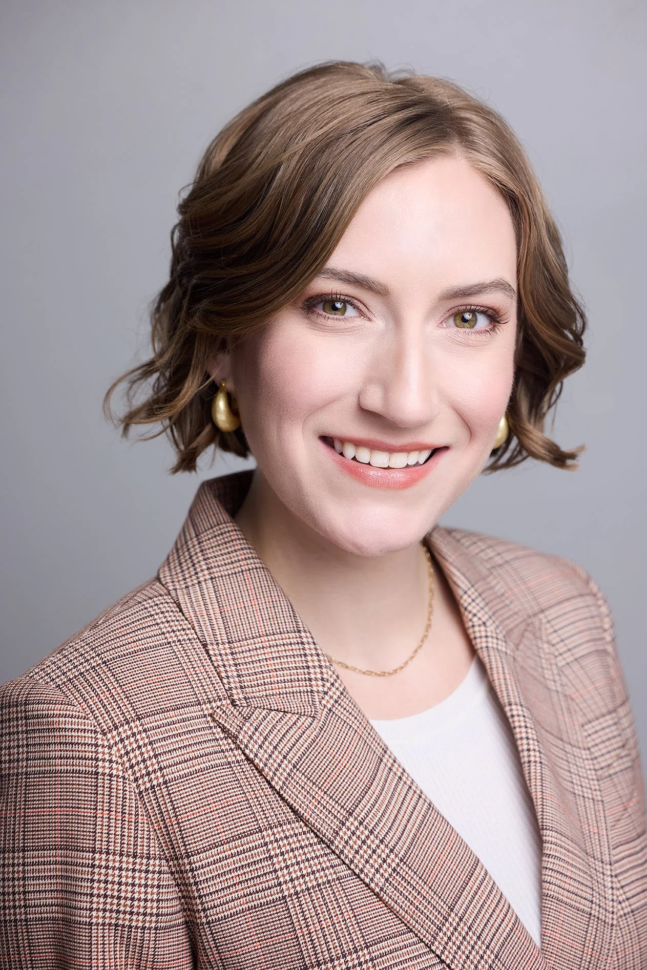 Headshot of young woman in plaid jacket smiling at camera on gray background.