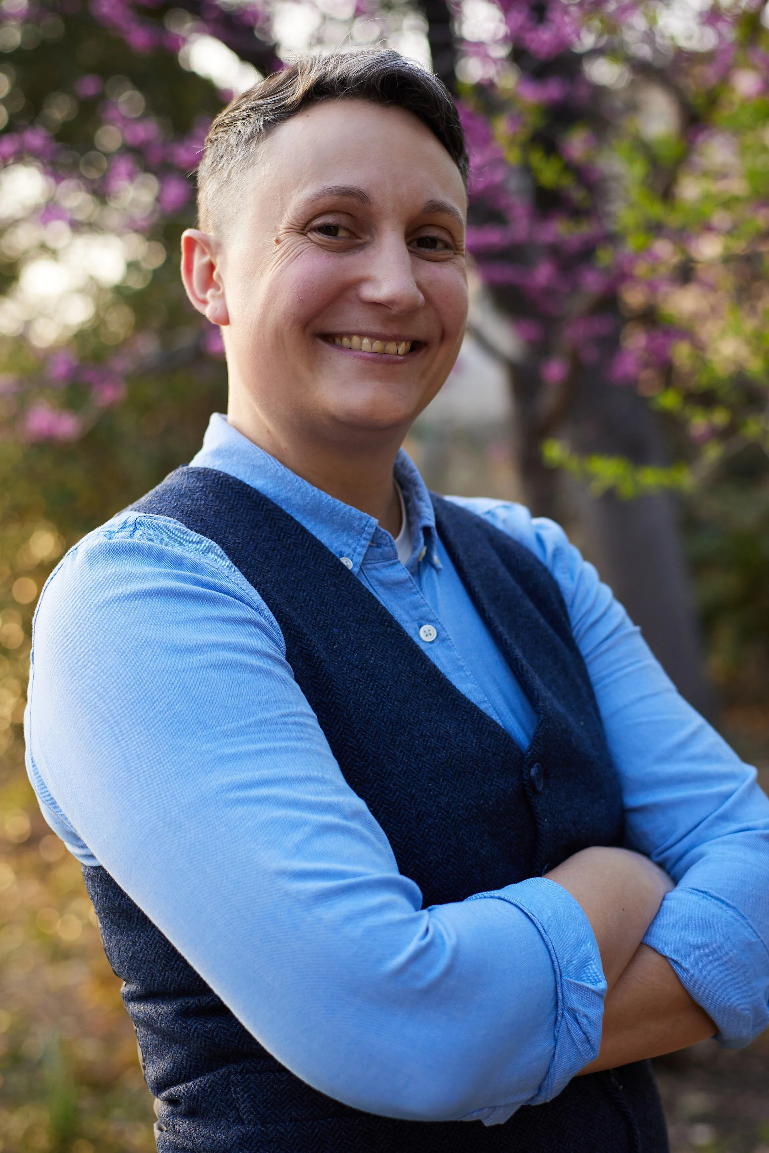 A smiling person with short hair standing outdoors in front of pink flowering trees, dressed in a blue shirt and a dark blue vest with arms crossed.