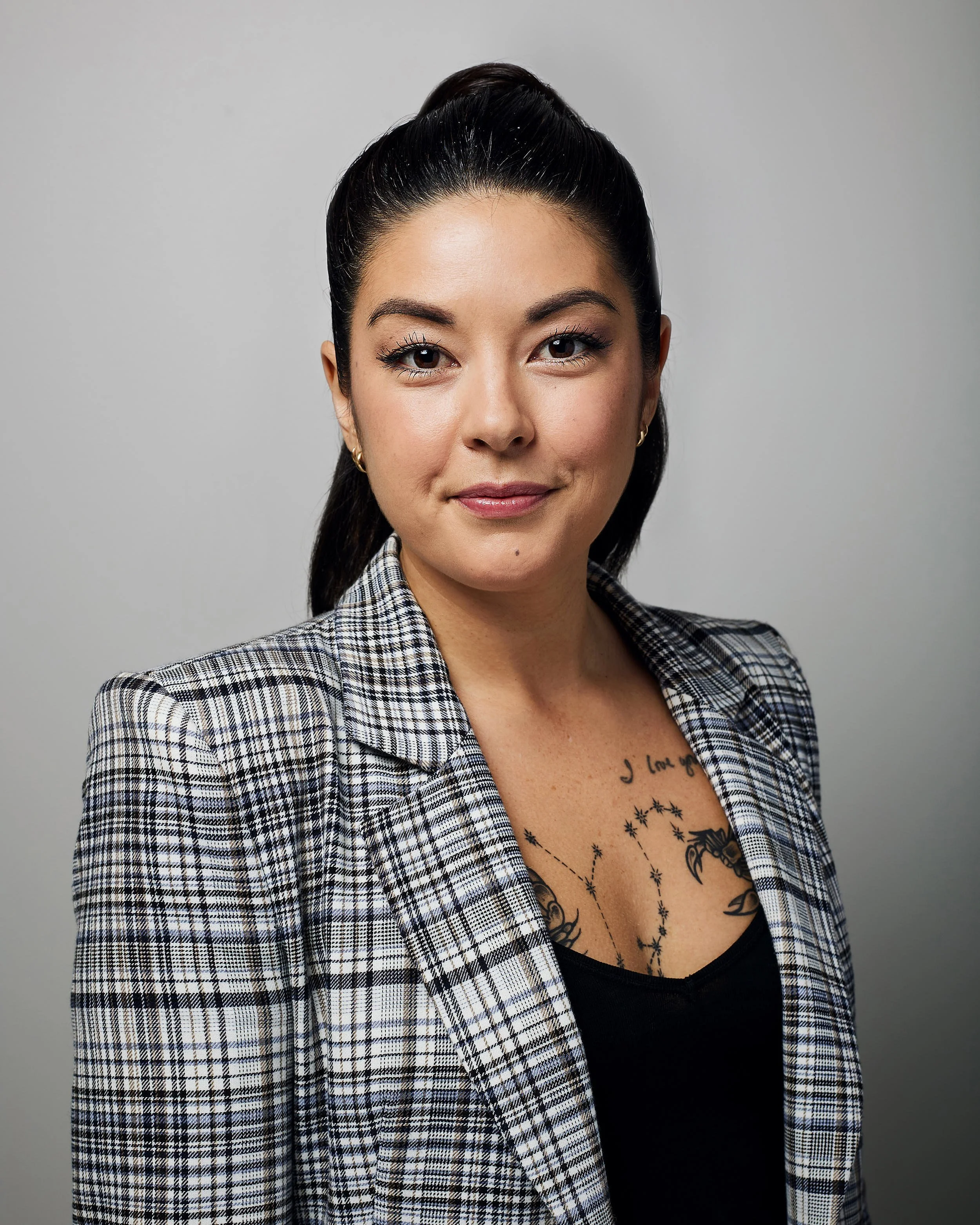 A woman with dark hair pulled back in a high ponytail, wearing a checked blazer and black top, standing against a plain background.