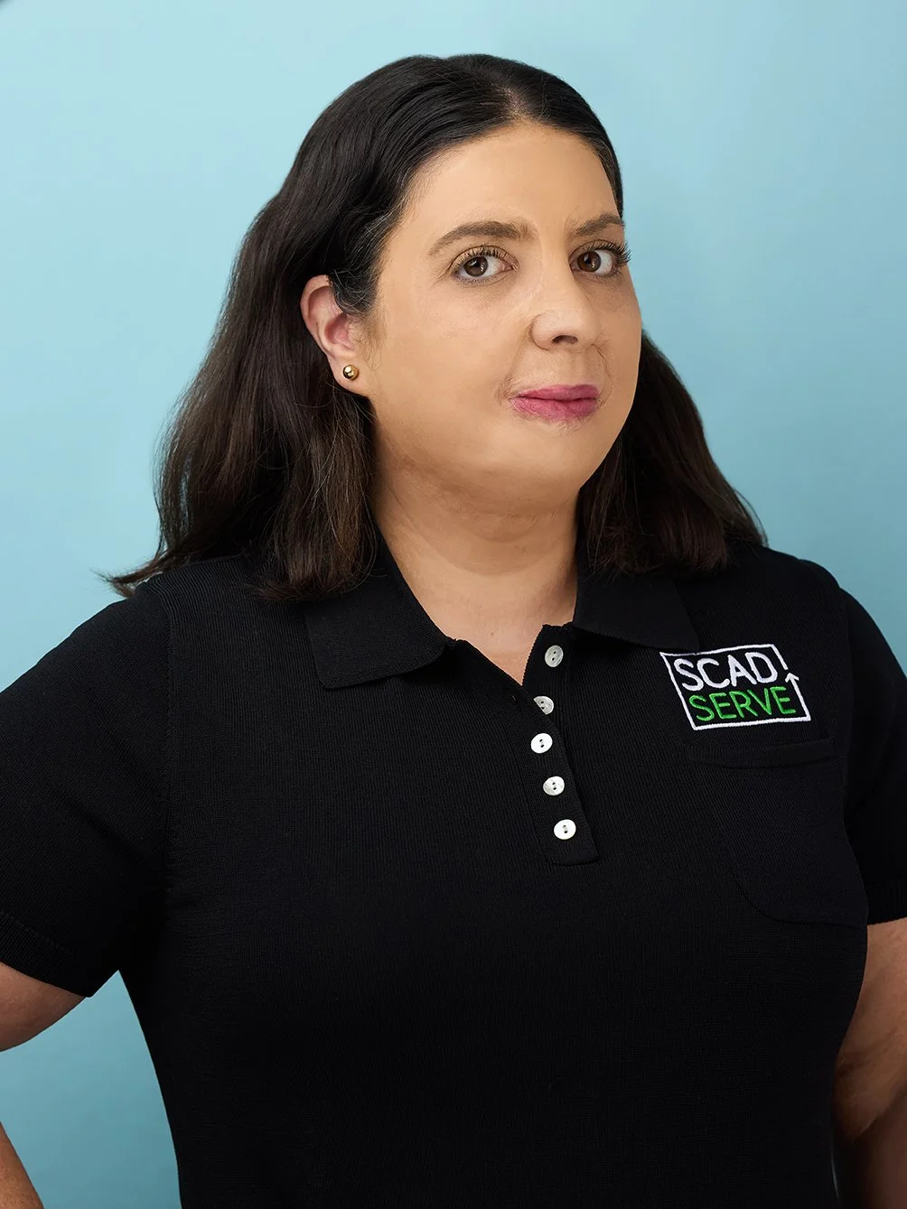 A woman with dark brown hair and fair skin wearing a black polo shirt with the logo 'SCAD SERVE' on it, standing against a light blue background.