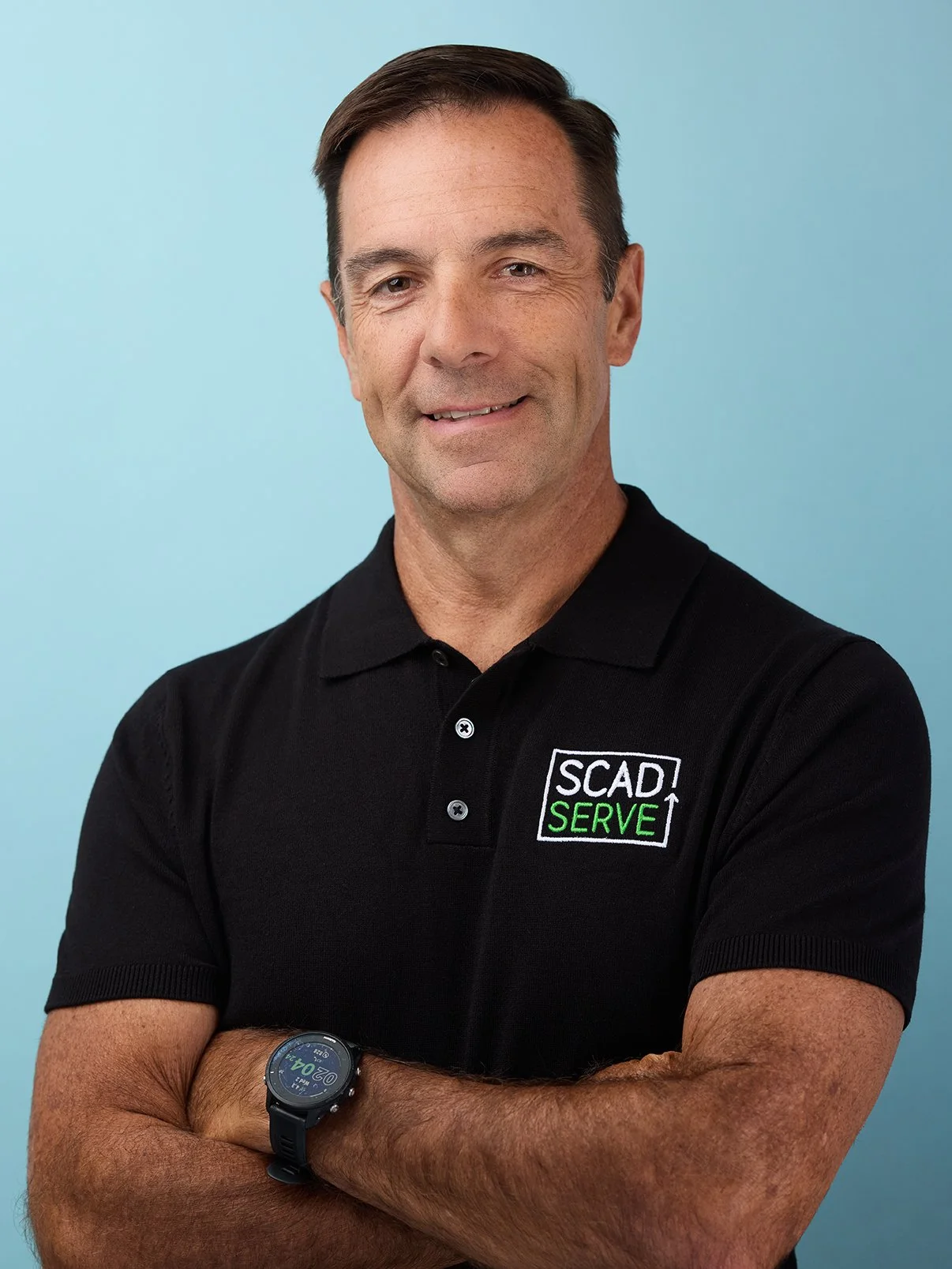 A man with short brown hair, wearing a black polo shirt with a logo that reads 'SCAD SERVE', standing with arms crossed against a light blue background.
