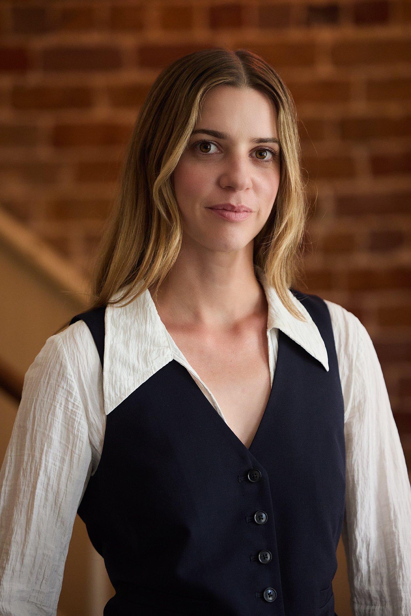A young woman with long, wavy light brown hair, wearing a white blouse and a black vest, standing indoors against a brick wall.