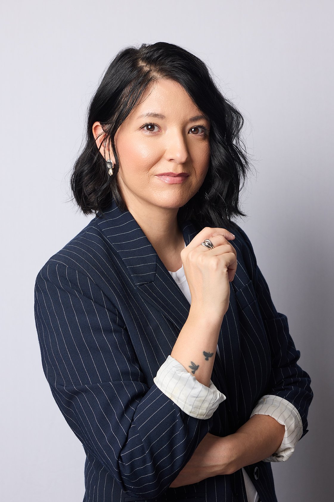 A woman with shoulder-length black hair, wearing a navy blue pinstripe blazer with rolled-up sleeves, standing against a plain light gray background, looking at the camera with a confident expression, with her left hand resting on her chin and her right arm crossed, showing a tattoo of three small birds on her inner wrist.