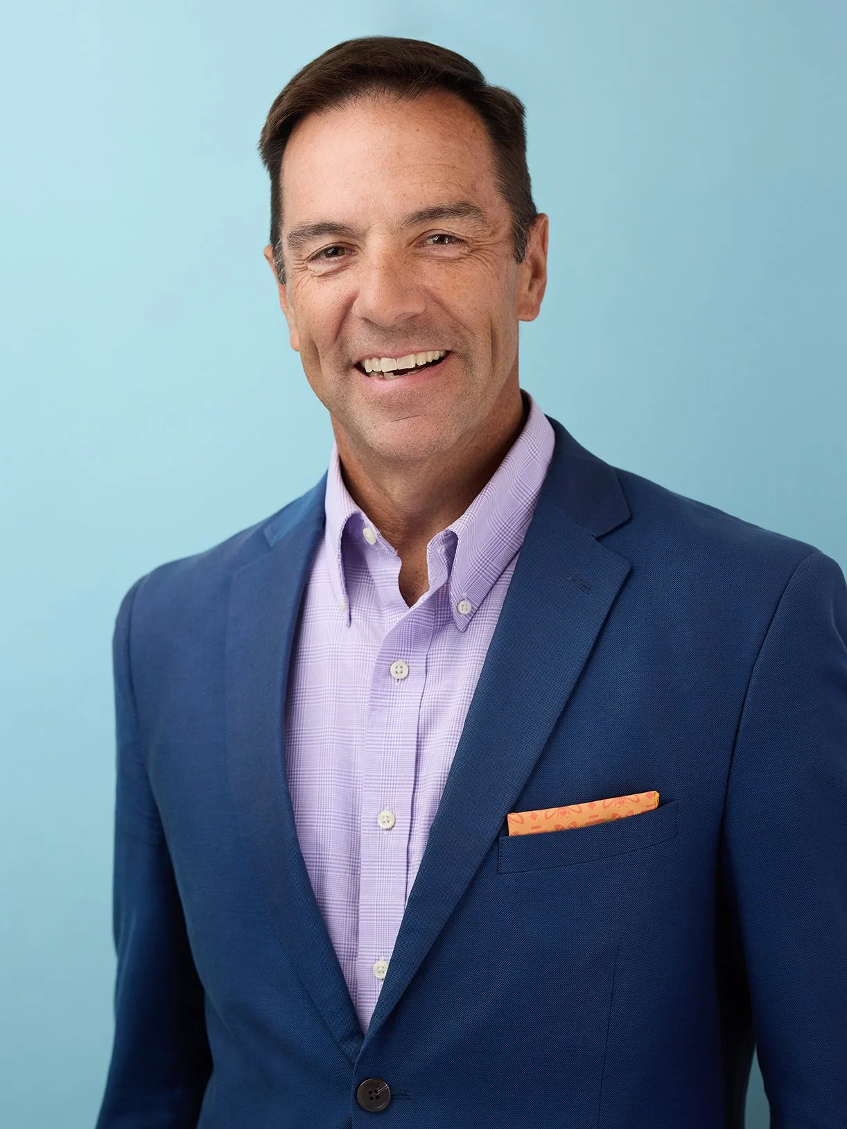 Smiling man in a blue suit jacket, light purple dress shirt, with a patterned pocket square, against a gradient blue background.
