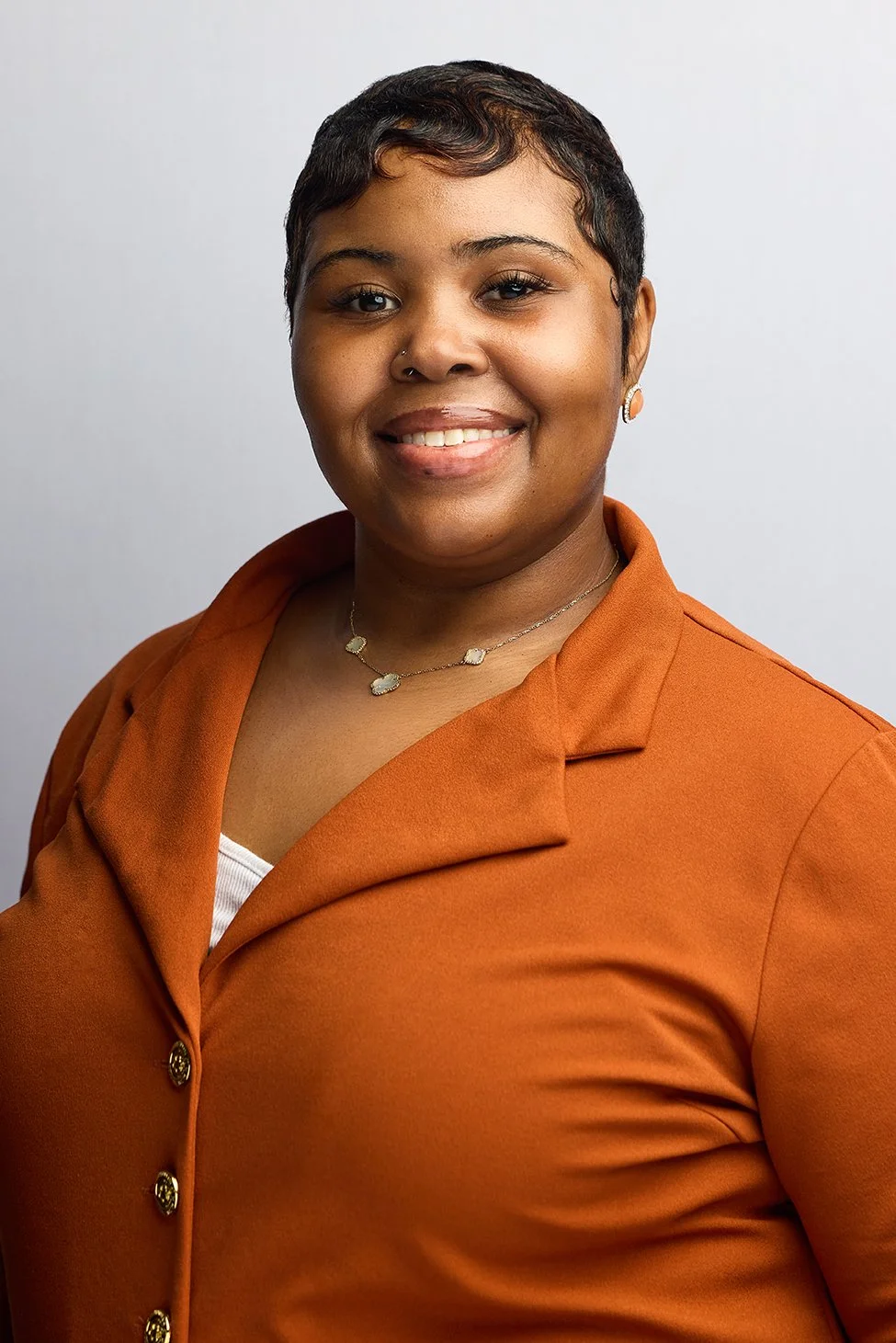 A woman with short, dark hair styled with waves, wearing an orange blazer over a white top, gold jewelry including a necklace and earrings, and smiling against a light gray background.