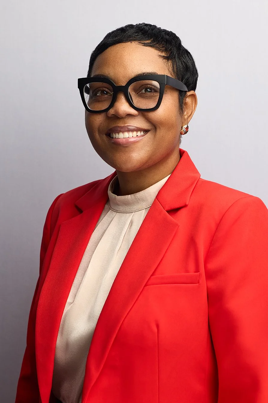 A woman with short black hair, wearing large black glasses, a red blazer, and a beige blouse, smiling against a plain light gray background.