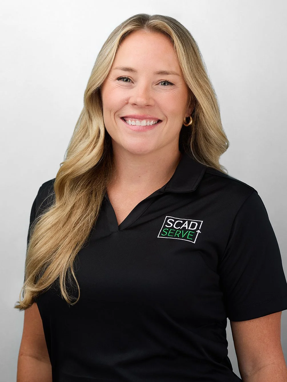 A woman with long blonde hair, smiling, wearing a black polo shirt with 'SCAD SERVE' logo, standing against a light gray background.