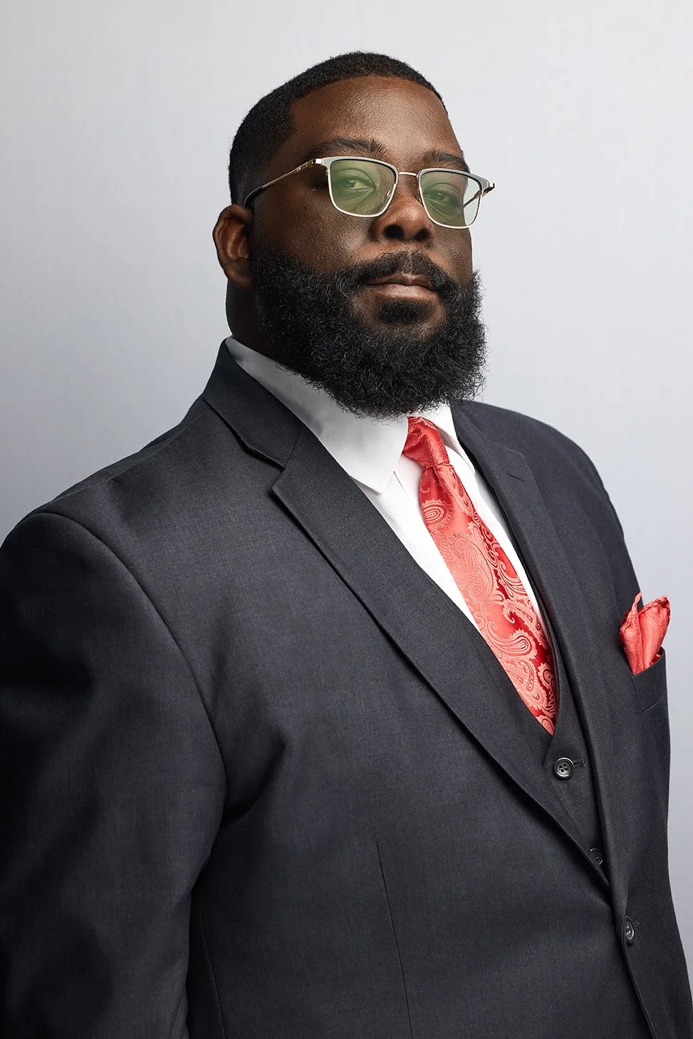 A man with a beard and glasses wearing a dark suit, white shirt, and red patterned tie and pocket square.