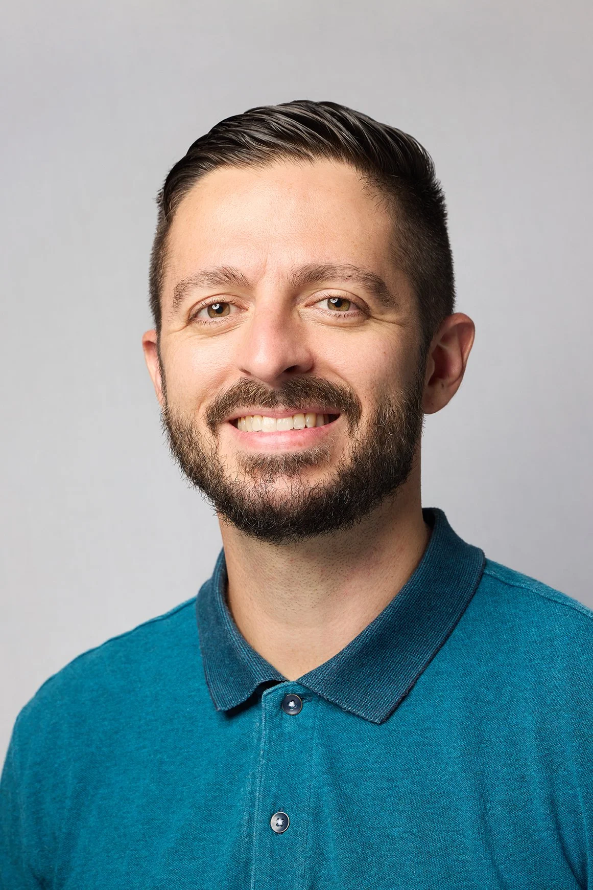 Headshot of smiling man with short dark hair, beard, wearing a blue button-up shirt, against a plain light gray background.