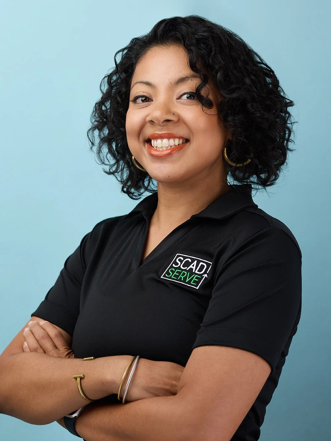 A woman with curly black hair smiling, wearing a black polo shirt with a patch that says 'SCAD Serve', standing against a light blue background.
