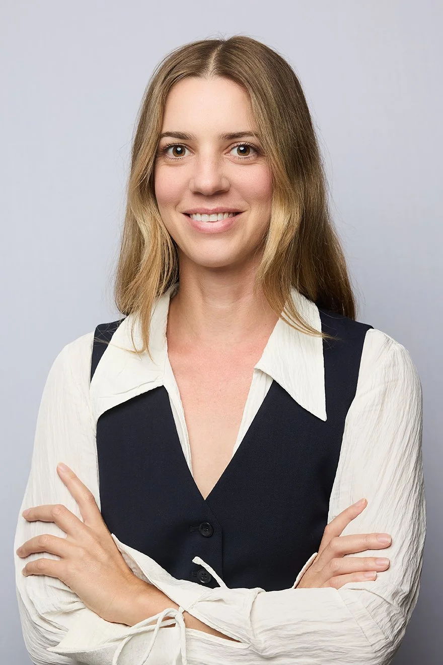 A young woman with light brown hair and a white blouse layered under a black vest, smiling with arms crossed, against a light grey background.