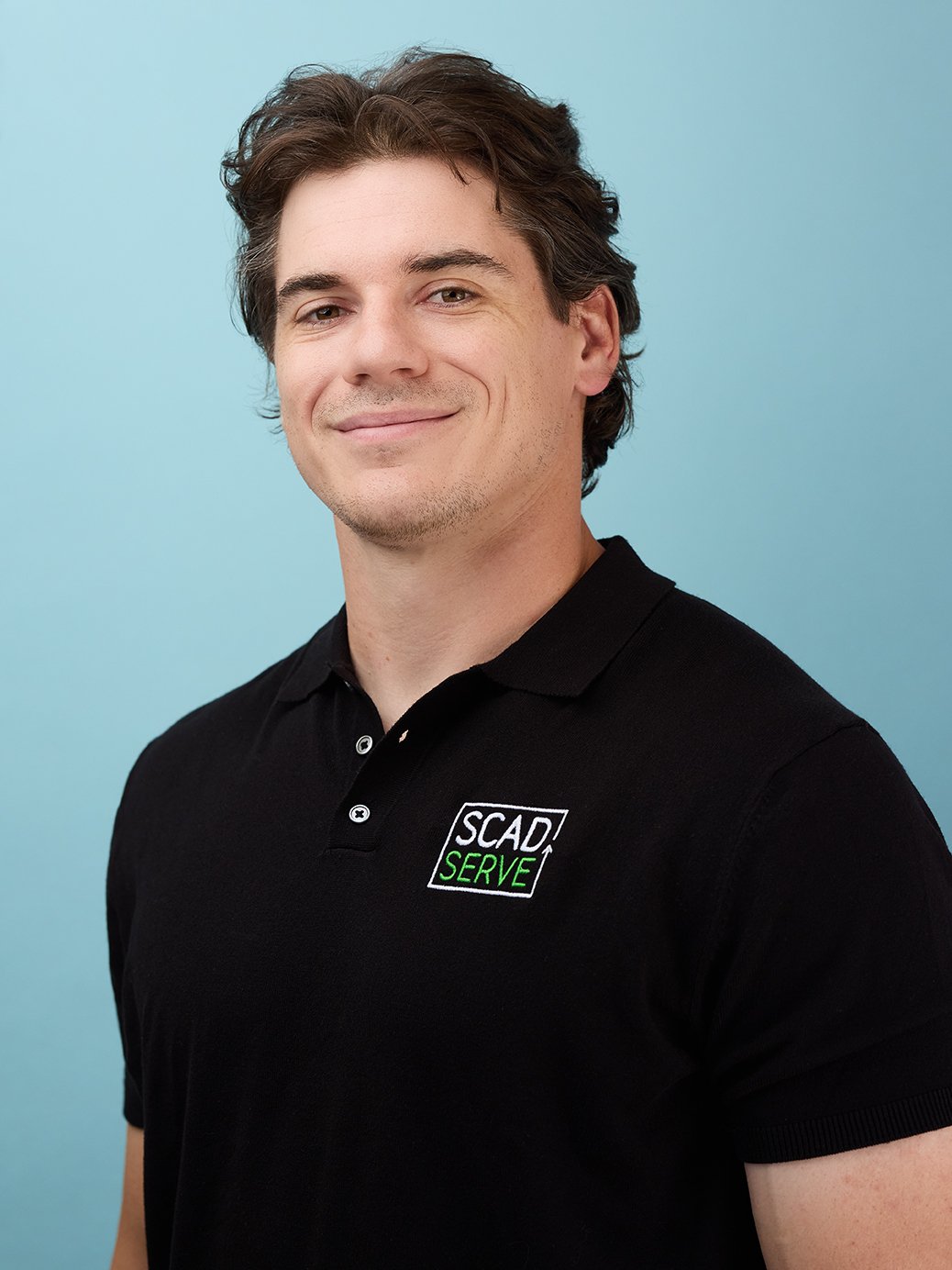 A young man with brown wavy hair smiling, wearing a black polo shirt with a logo that says 'SCAD Serve' on the chest, standing against a light blue background.