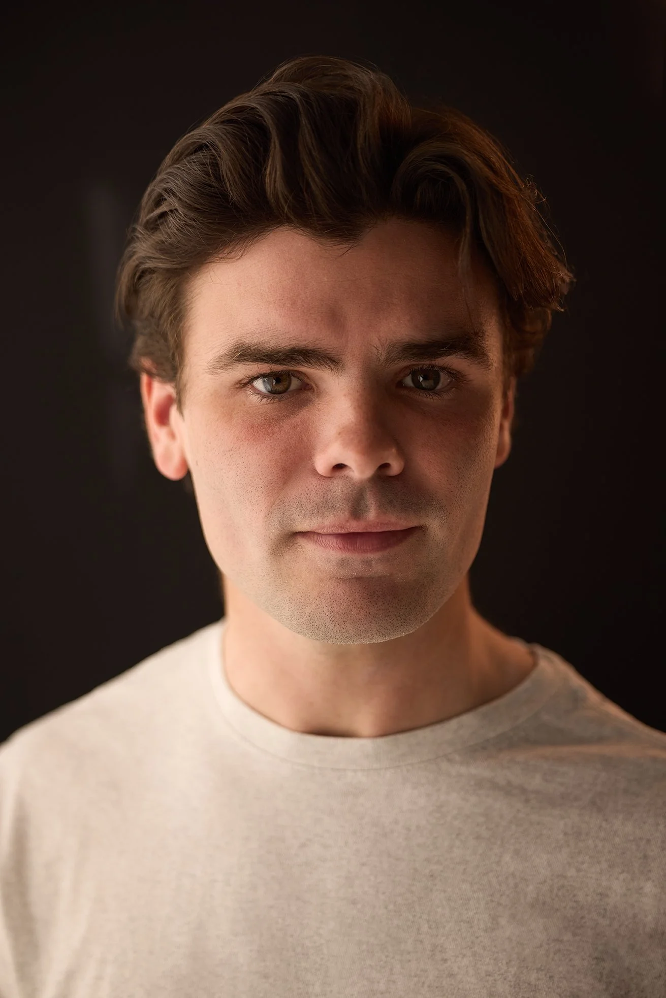 Close-up portrait of a young man with brown hair and light skin, wearing a beige shirt, against a dark background.