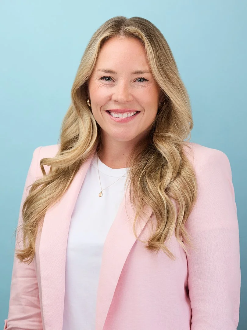 A woman with long blonde hair, wearing a pink blazer over a white top, smiling against a light blue background.