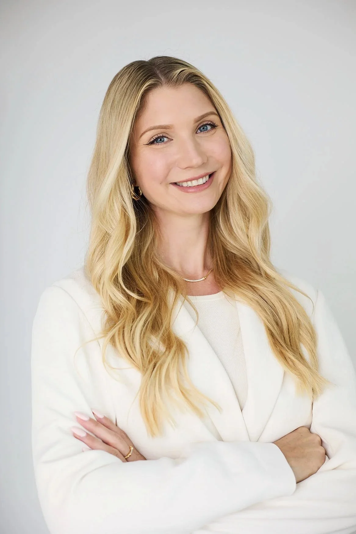 A woman with long blonde hair and blue eyes smiling, wearing a white blazer and gold jewelry, standing against a plain white background.