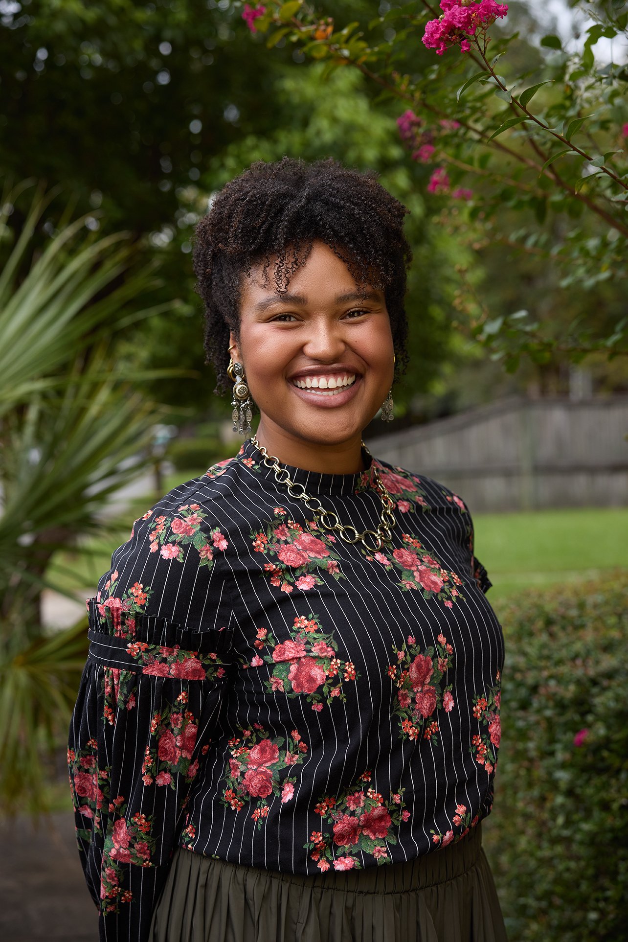 A woman smiling outdoors with greenery and pink flowers in the background, wearing a black floral blouse, earrings, and a necklace.