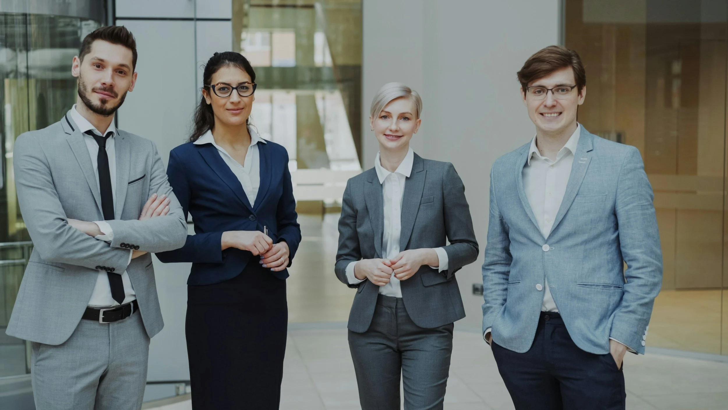 Four professionally dressed people standing together in an office building, smiling at the camera.