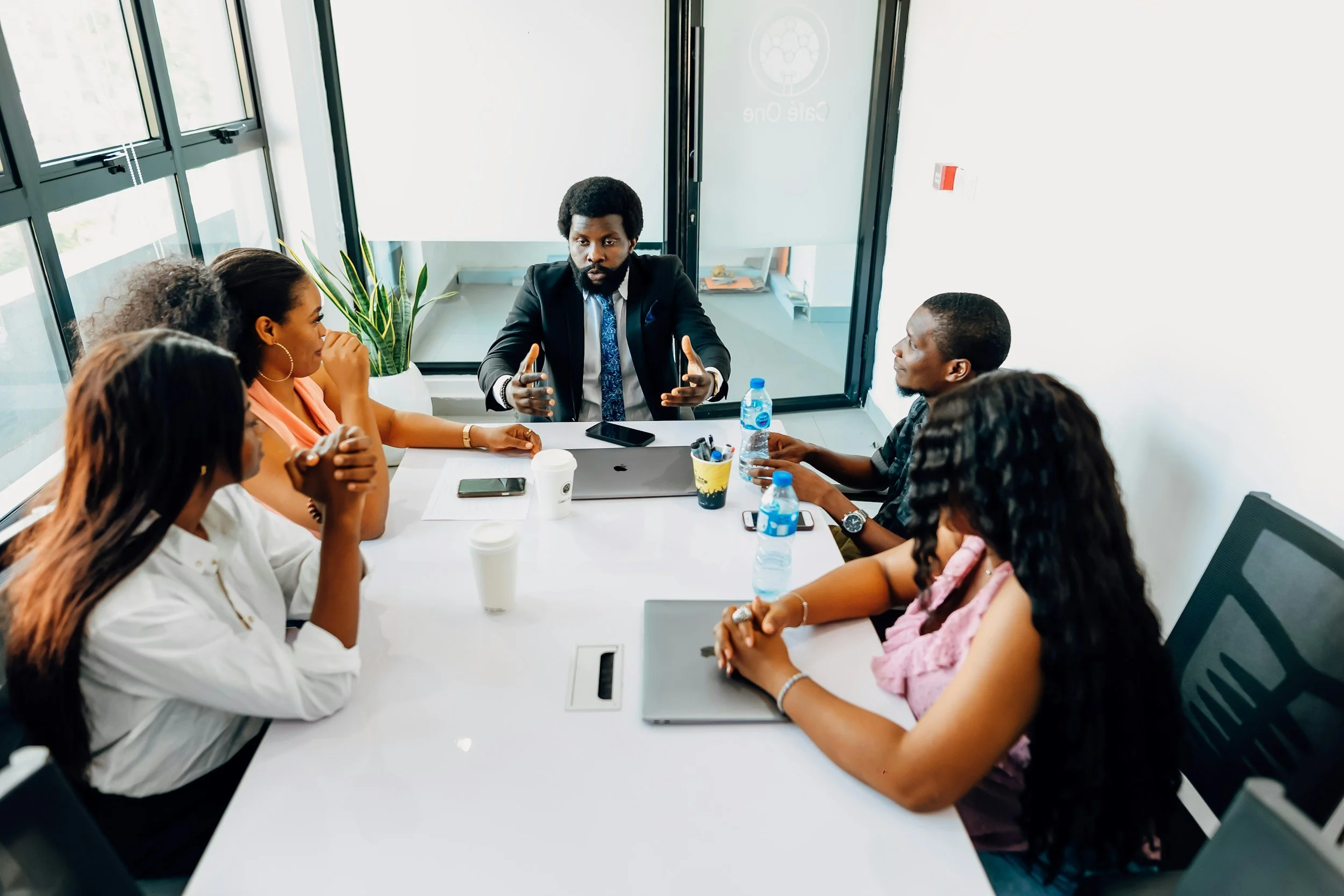 A man in a suit leads a business meeting with five women around a white conference table in a modern office, with laptops, water bottles, and coffee cups on the table.