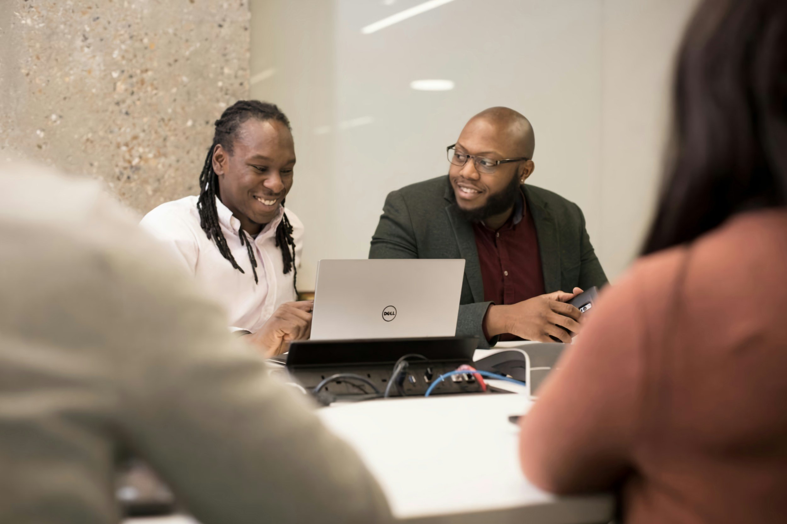 Two men and two women in a meeting room, engaging in a discussion, with laptops and electronic devices on the table.