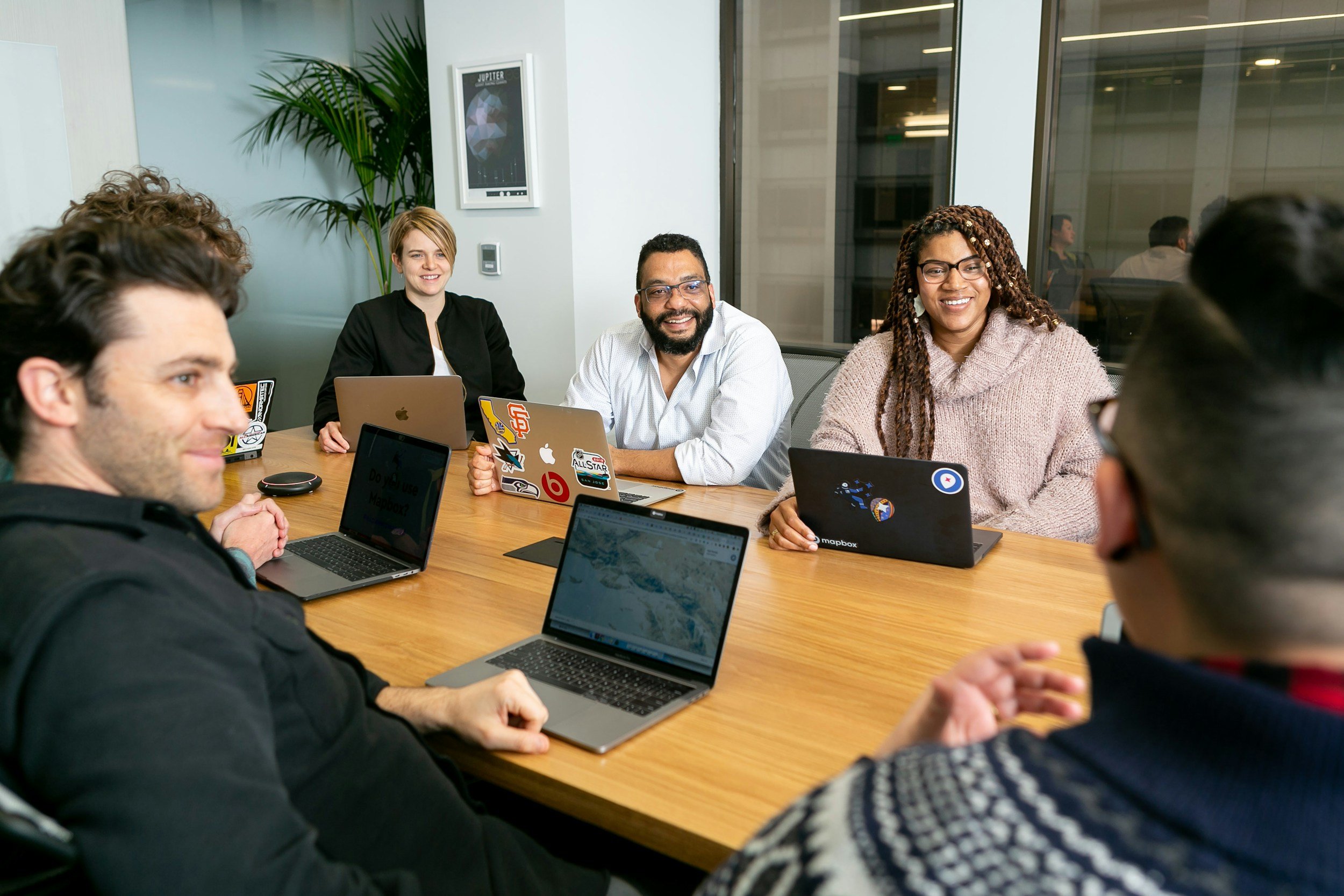 A diverse group of six people sitting around a conference table in a modern office, engaged in a meeting. They are using laptops, some with stickers, and smiling. There is a large window and a plant in the background.