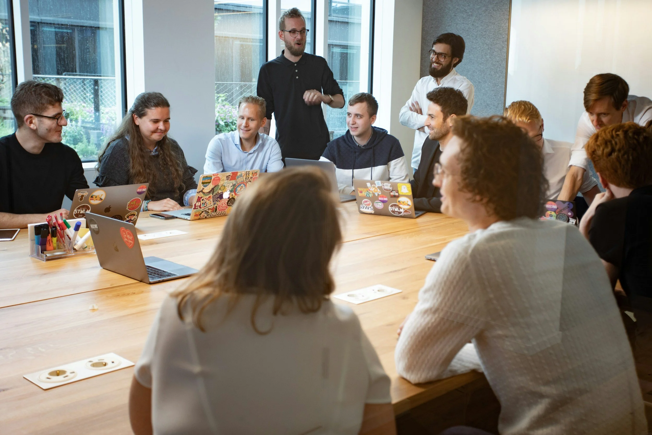 A group of diverse people gathered around a conference table during a meeting, with four individuals standing at the end of the table and others seated, all smiling and engaged.