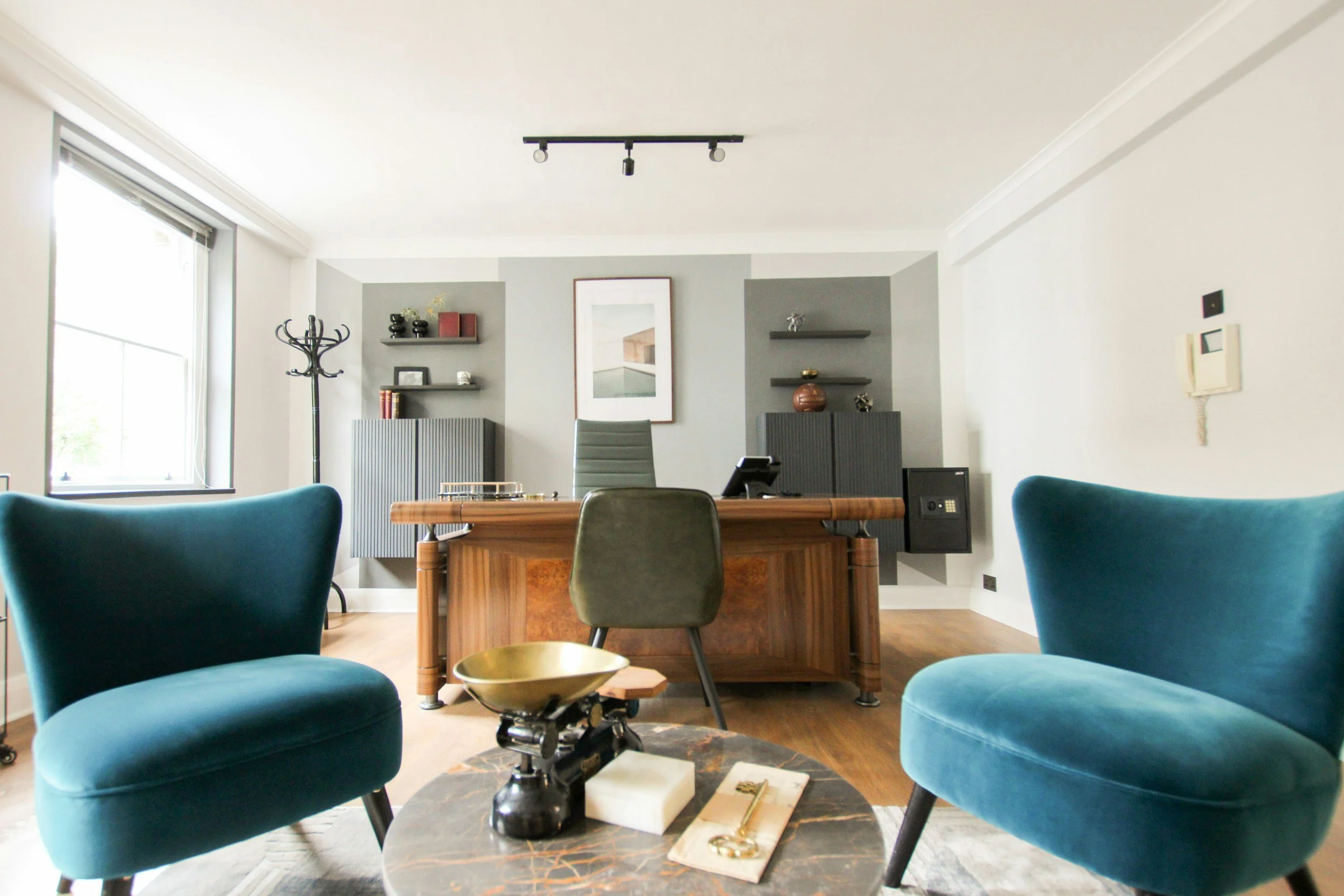 Modern office with two teal velvet armchairs in the foreground, a wooden desk with a gray office chair in the background, and shelves with decorative objects on a gray accent wall.