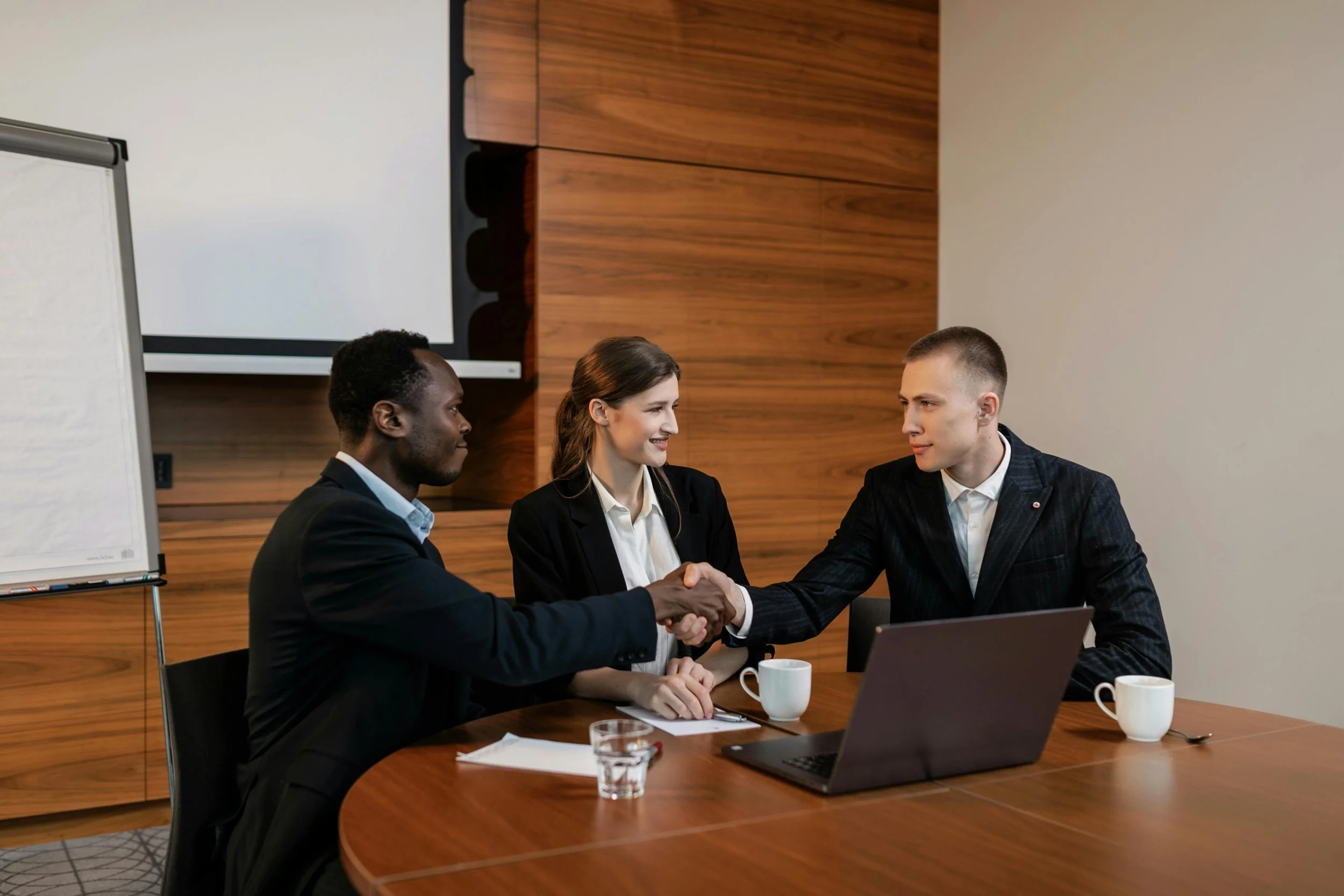 Three business professionals, two men and one woman, sitting at a conference table in a modern office, shaking hands, with laptops, cups, and documents on the table.