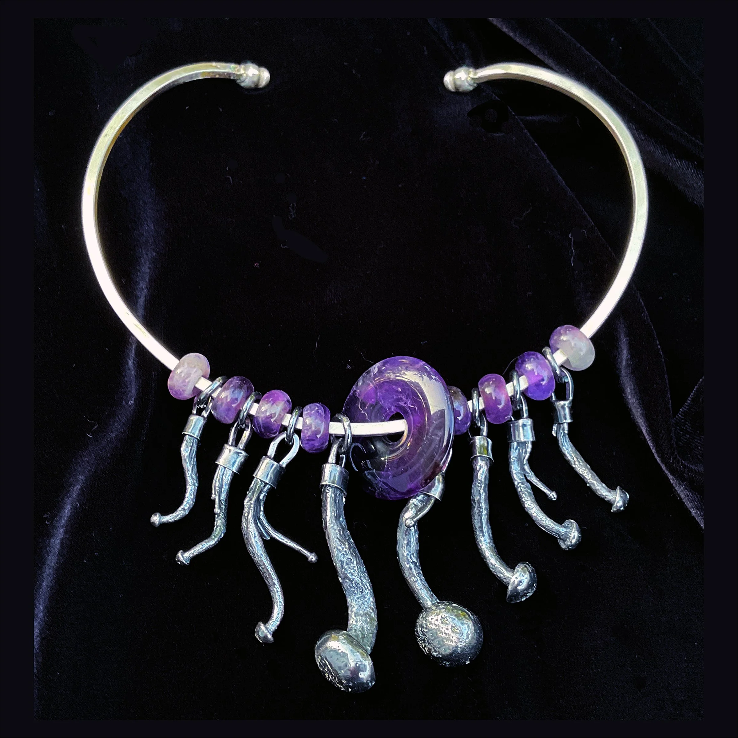 A silver choker necklace with amethyst beads, a large purple amethyst pendant, and silver mushroom charms hanging from it, placed on black fabric.