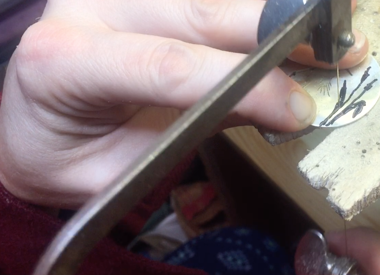 Close-up of a person using a jeweler's saw to cut a small round object with a black floral pattern.