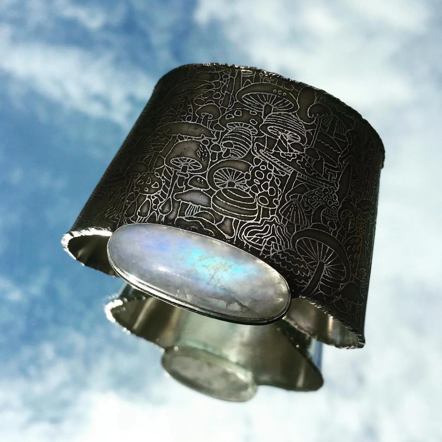 A sterling cuff with intricate etched mushroom designs featuring a moonstone viewed against a blue sky with clouds.