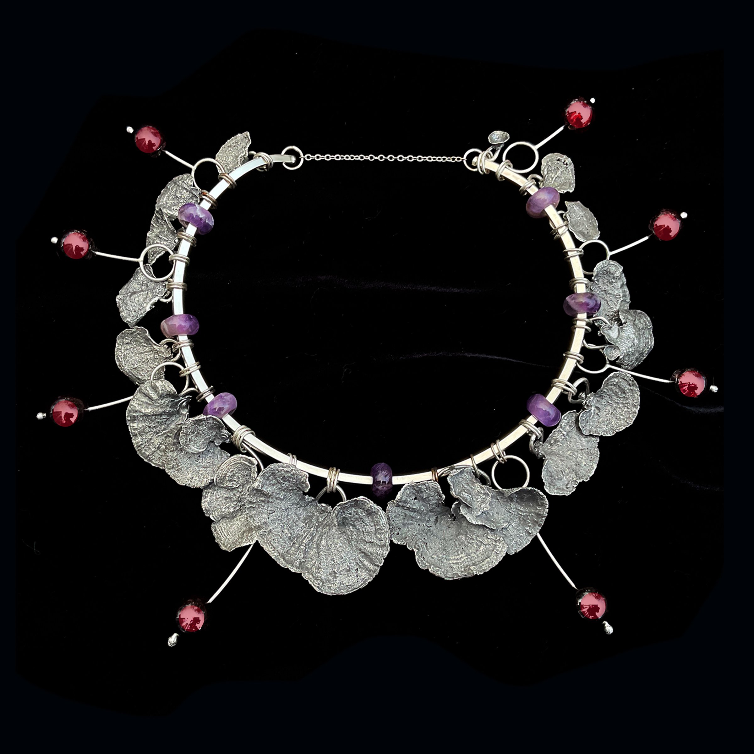 Silver necklace with textured silver leaves, round purple beads, and dangling red beads on silver wires, displayed on black fabric.