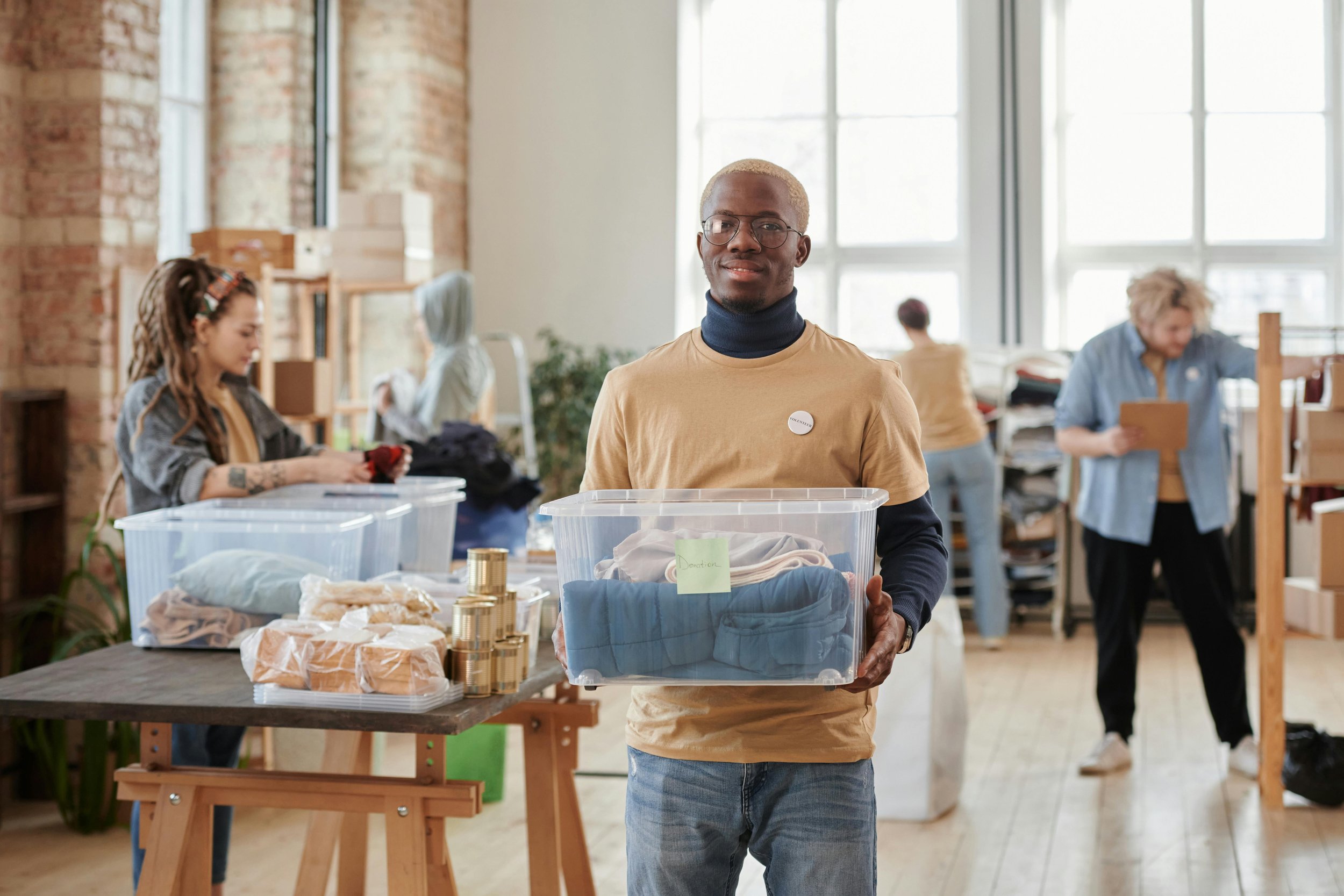 A man with glasses, blonde hair, and a beige T-shirt holding a clear plastic container filled with donated clothes inside a room with large windows and brick walls, with other people sorting and organizing items in the background.
