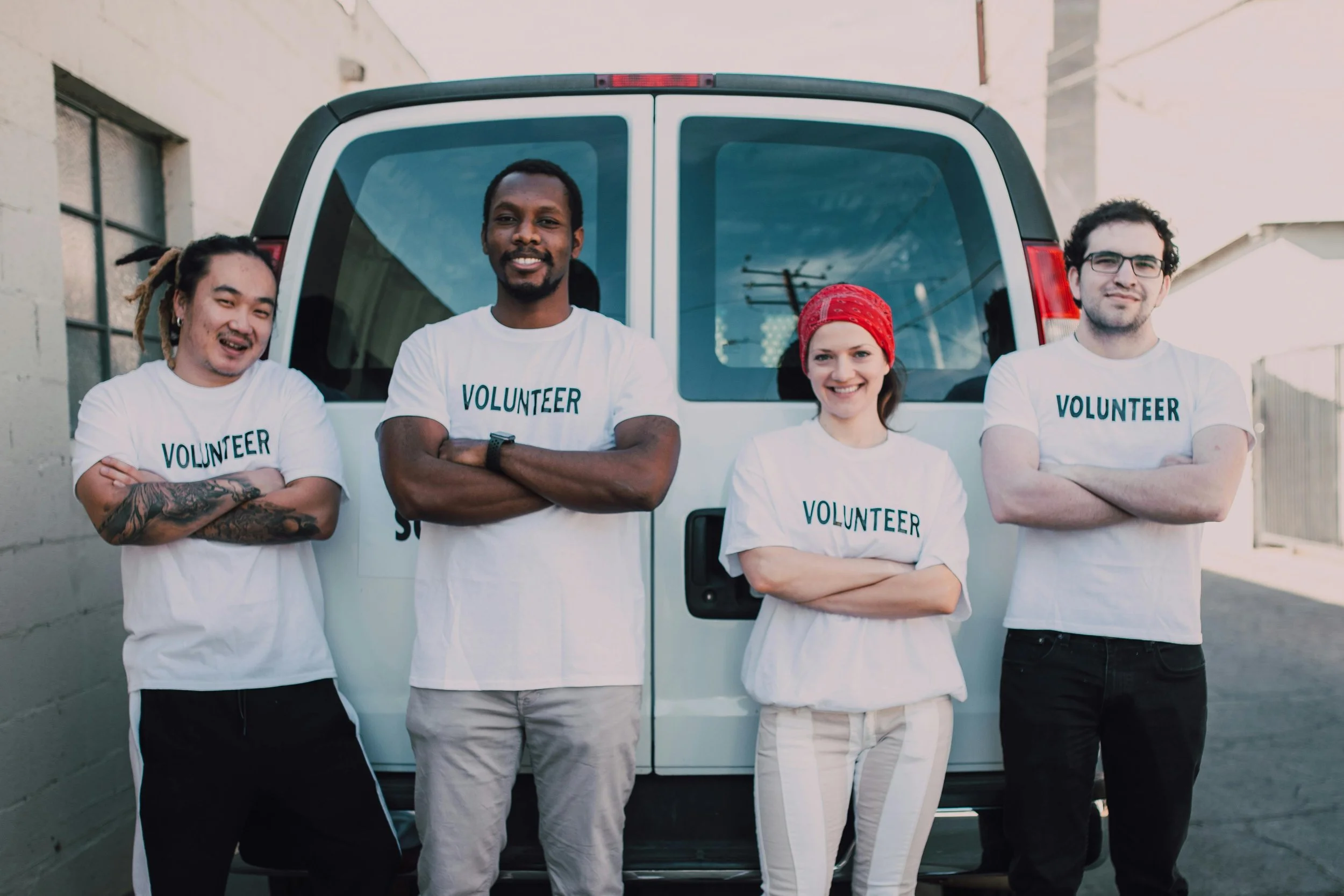 Four diverse volunteers standing in front of a white van, smiling with arms crossed, wearing white T-shirts labeled 'VOLUNTEER' in an urban setting.