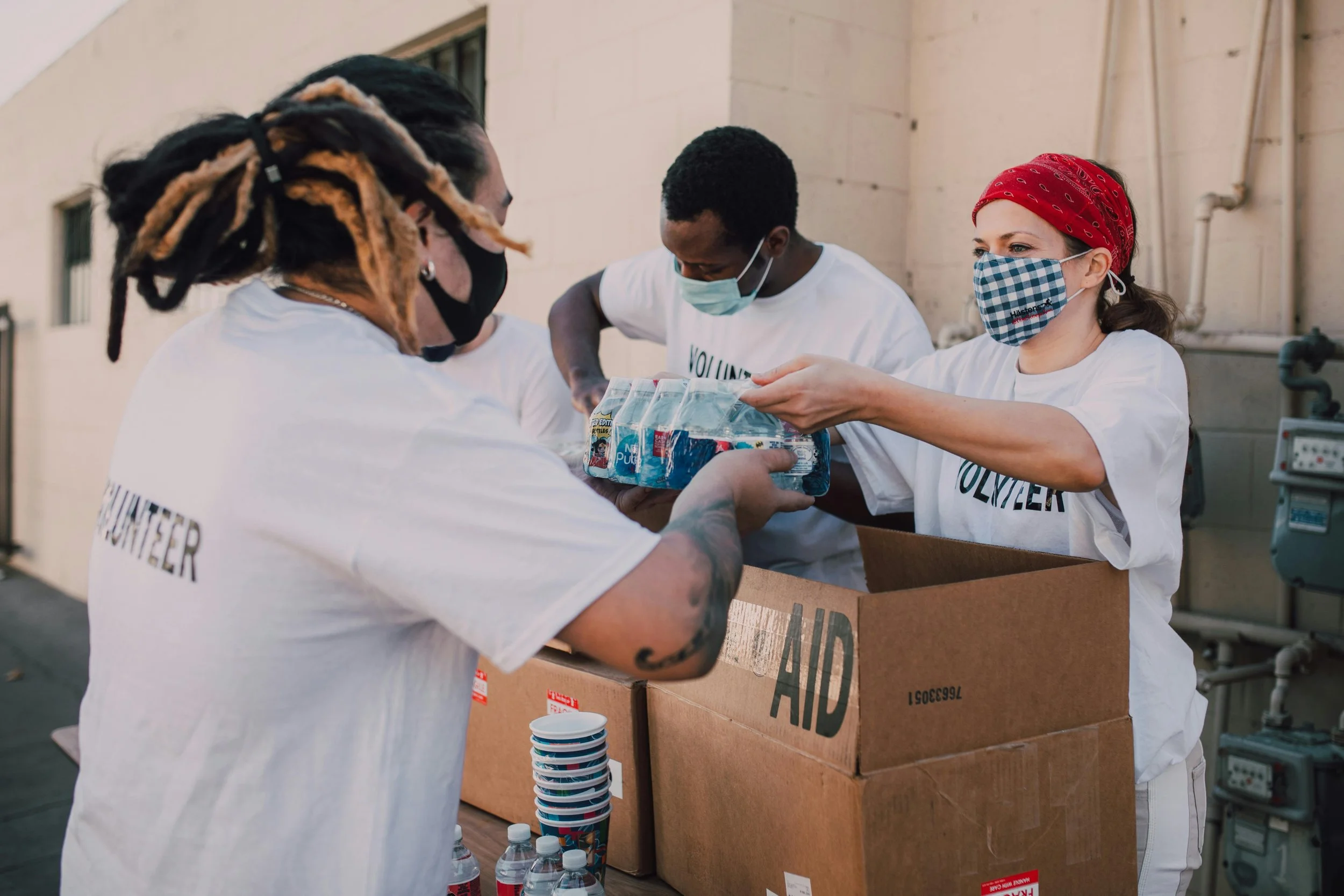 Four volunteers wearing face masks and white t-shirts packing bottled water and supplies into boxes labeled "AID" outdoors.