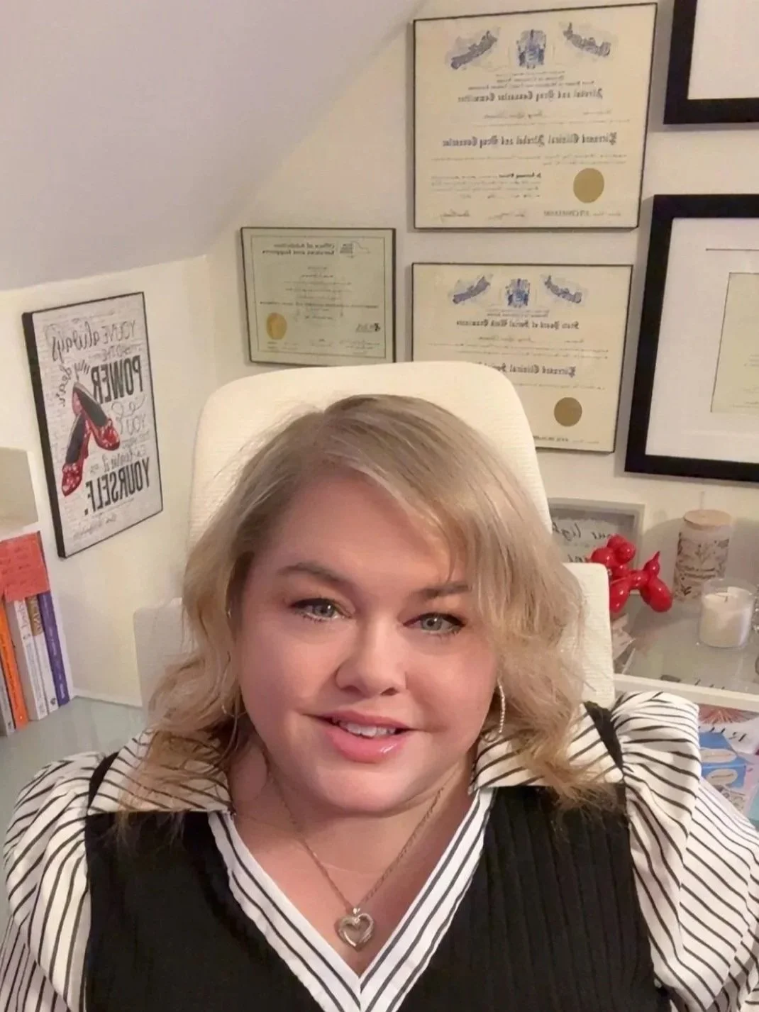 A woman with blonde, curly hair and hoop earrings takes a selfie while sitting in an office or study. Behind her on the wall are framed certificates, a framed quote poster, and other framed documents. A white desk with books, a red balloon animal, and various papers are visible.