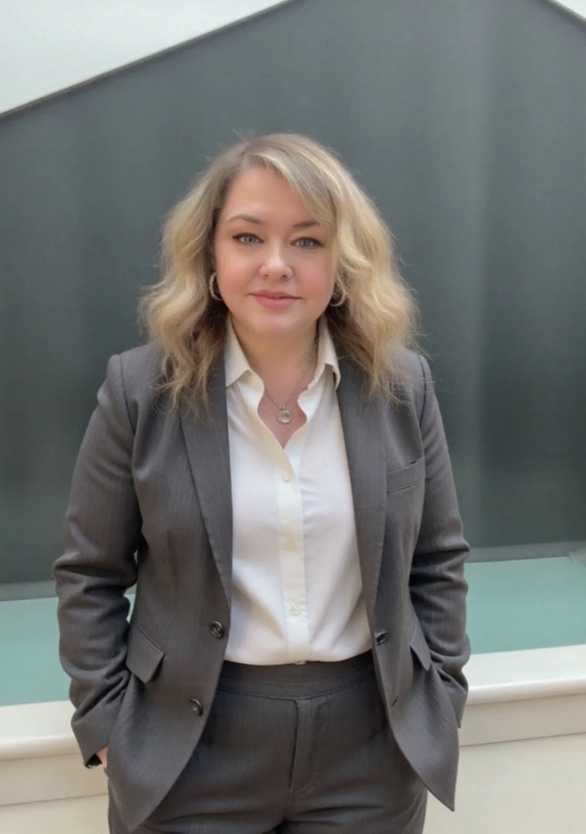 A woman with blonde, wavy hair wearing a dark gray business suit and white shirt standing in front of a glass wall.